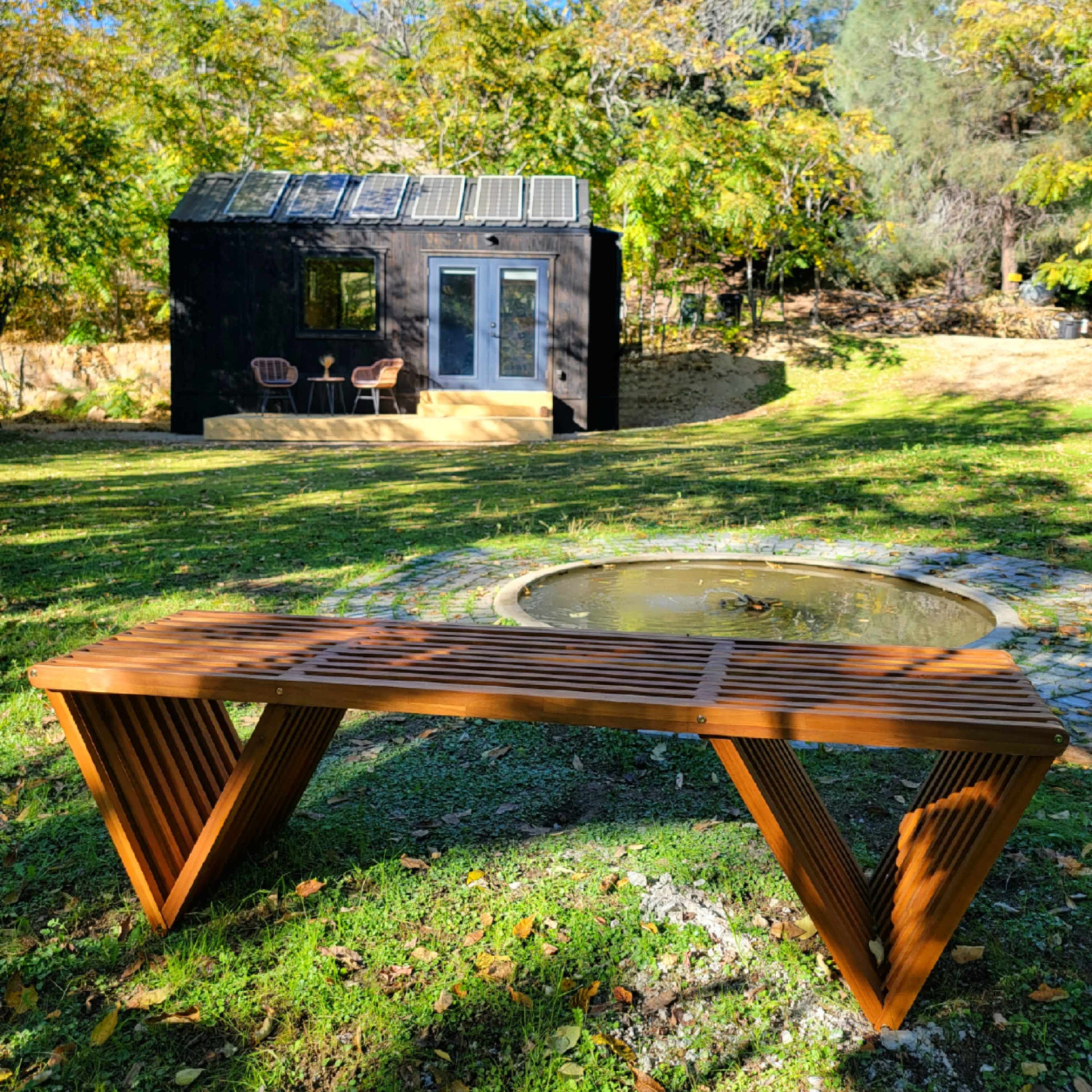 A wooden bench is positioned in front of a small black cabin surrounded by greenery and trees.