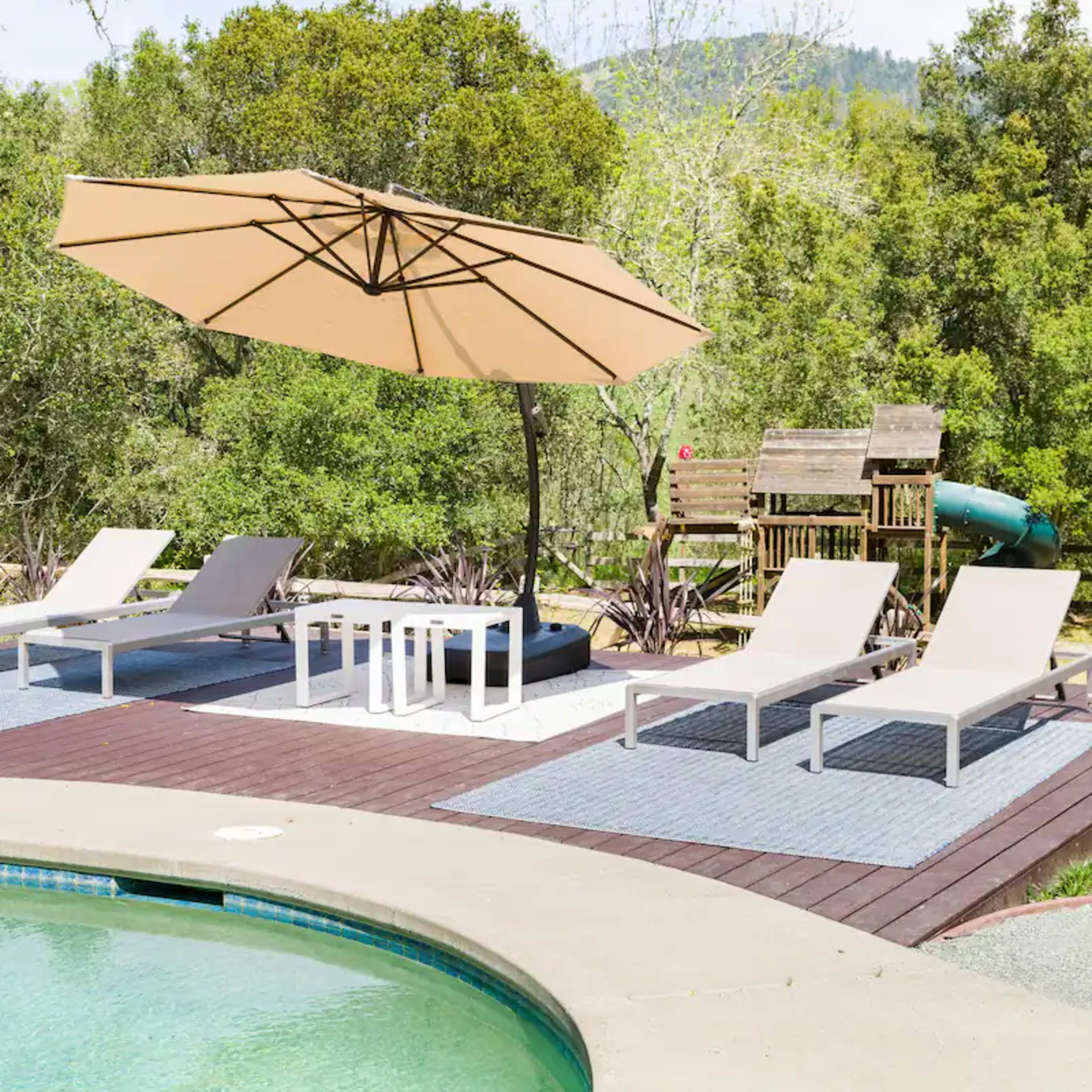 A pool area features two lounge chairs, a table, and a large umbrella on wooden decking beside the water, surrounded by greenery.