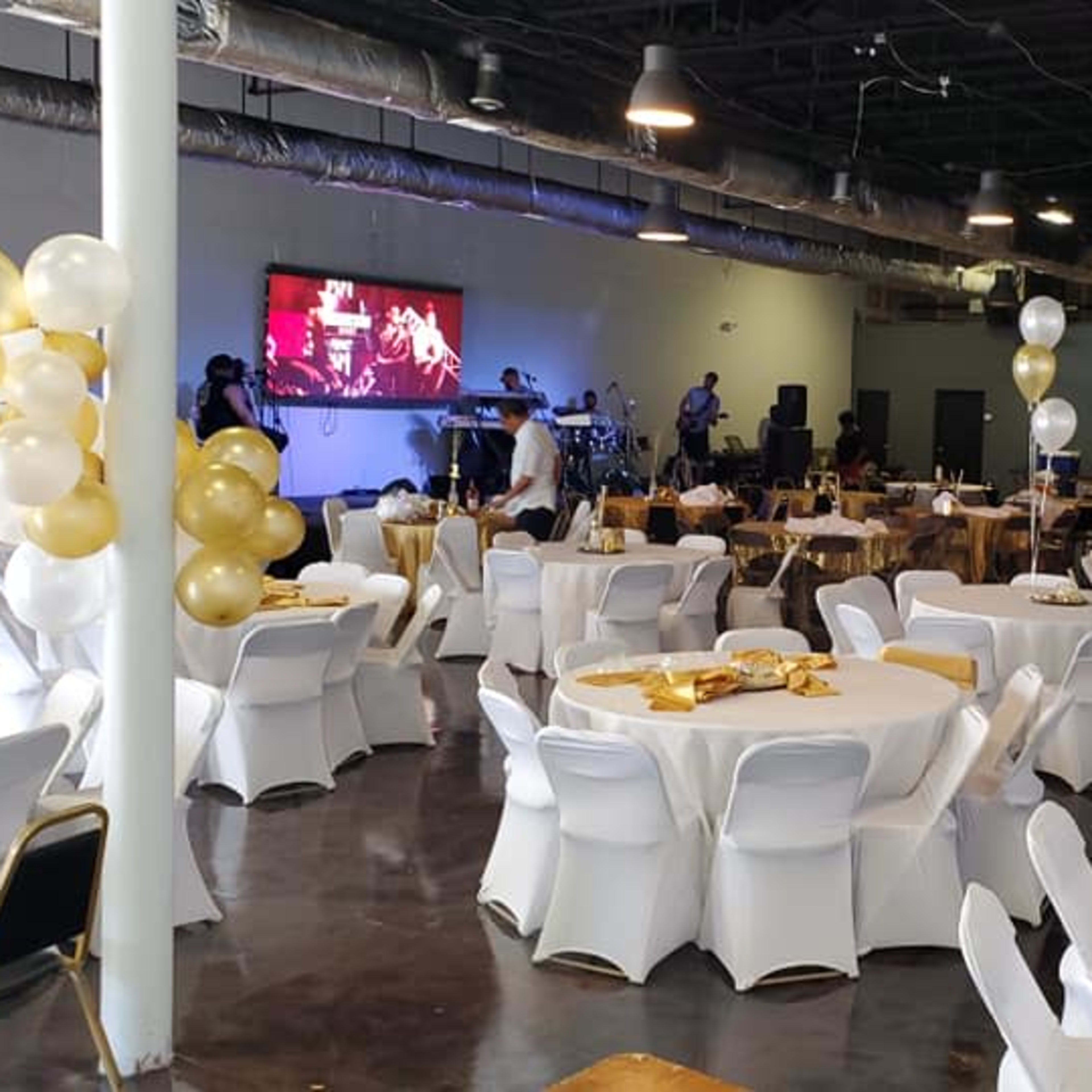 A banquet hall set up for an event, featuring round tables with white tablecloths, gold decorations, and a stage in the background with a band.