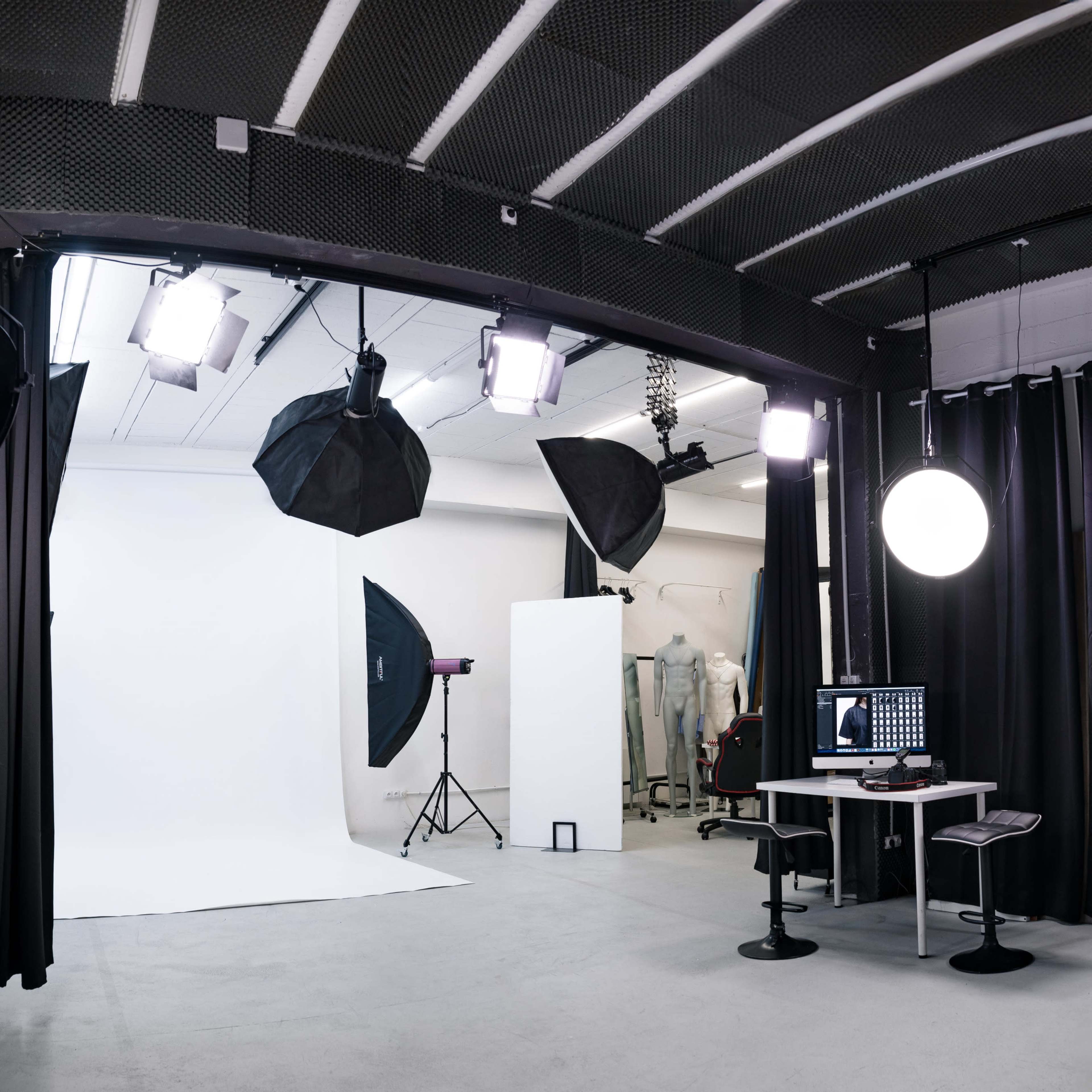 A photography studio features a white backdrop, multiple light sources, a computer setup, and dark curtains.