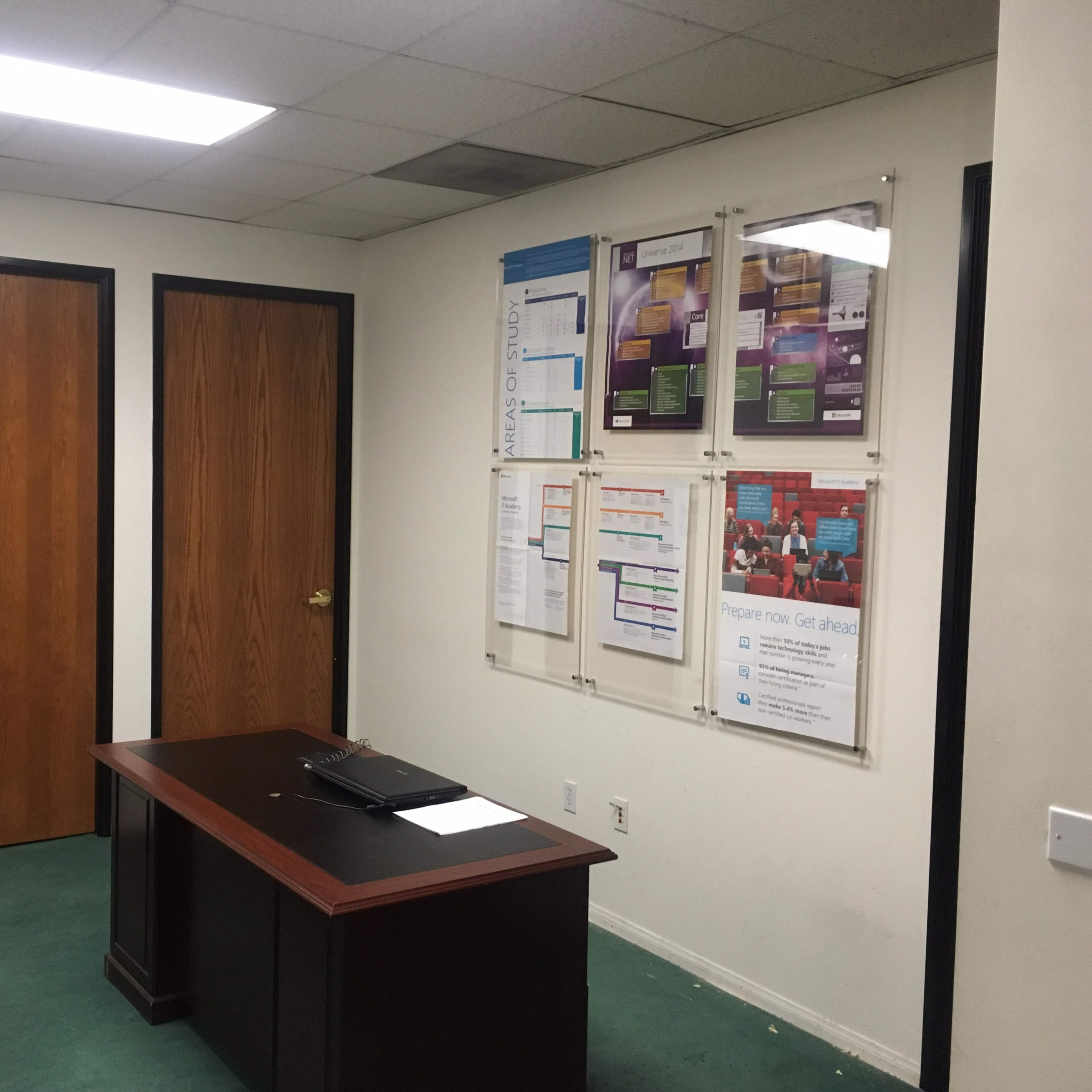 A small office space with two closed wooden doors, a wooden desk, and information posters mounted on the wall.