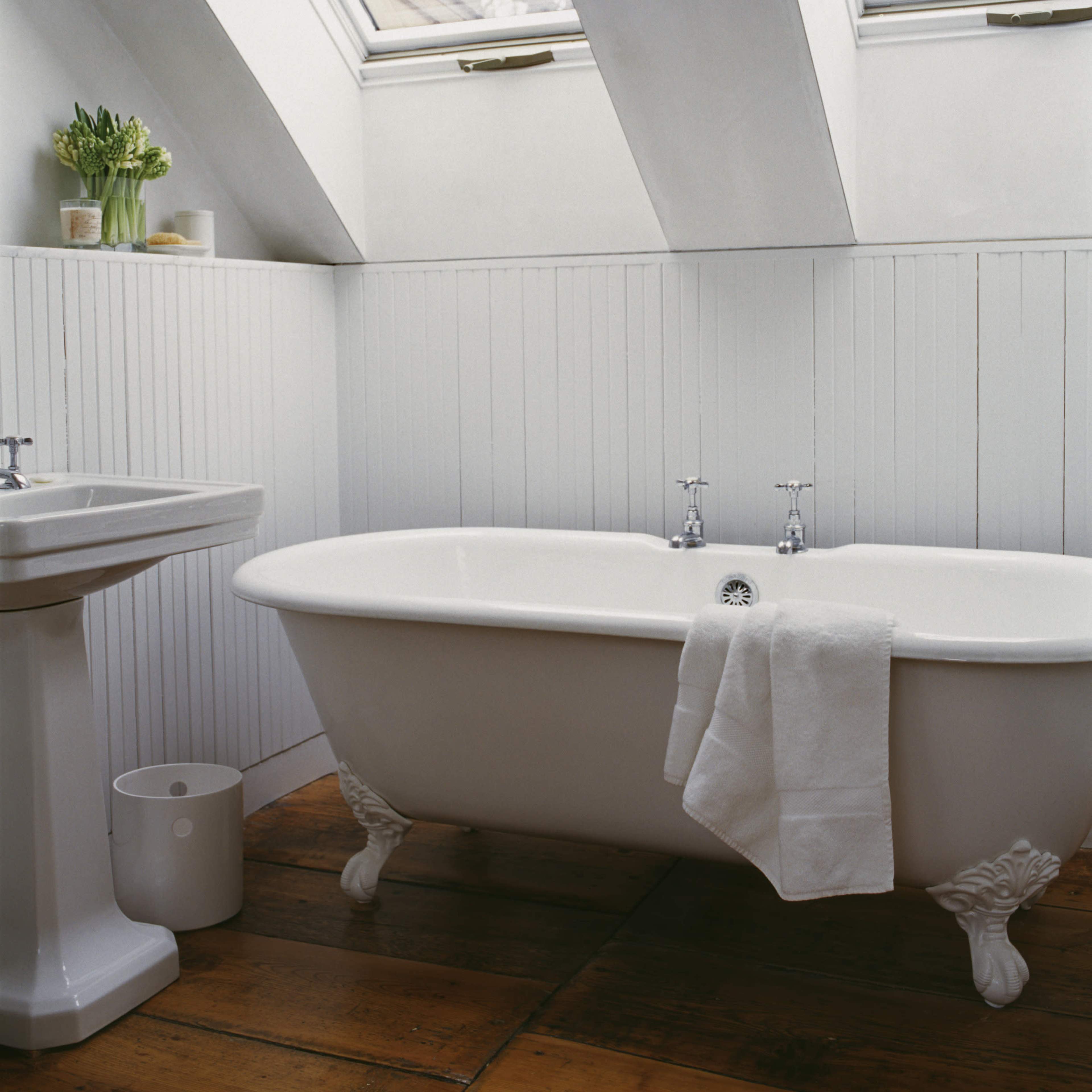 A white clawfoot bathtub is positioned beneath slanted skylights in a minimalist bathroom with wooden flooring and a sink nearby.