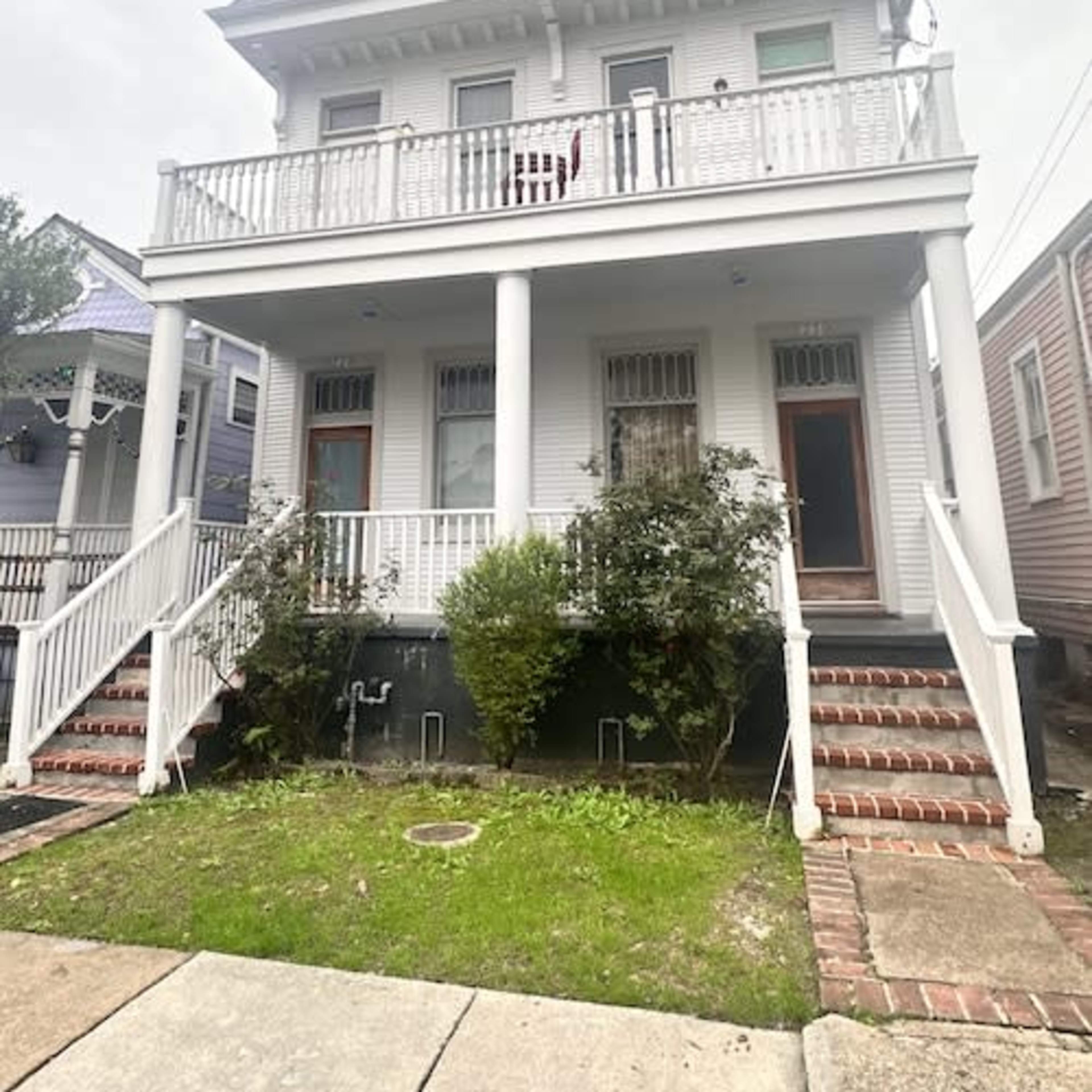 A two-story white house with a balcony and a front staircase stands among other similar homes.