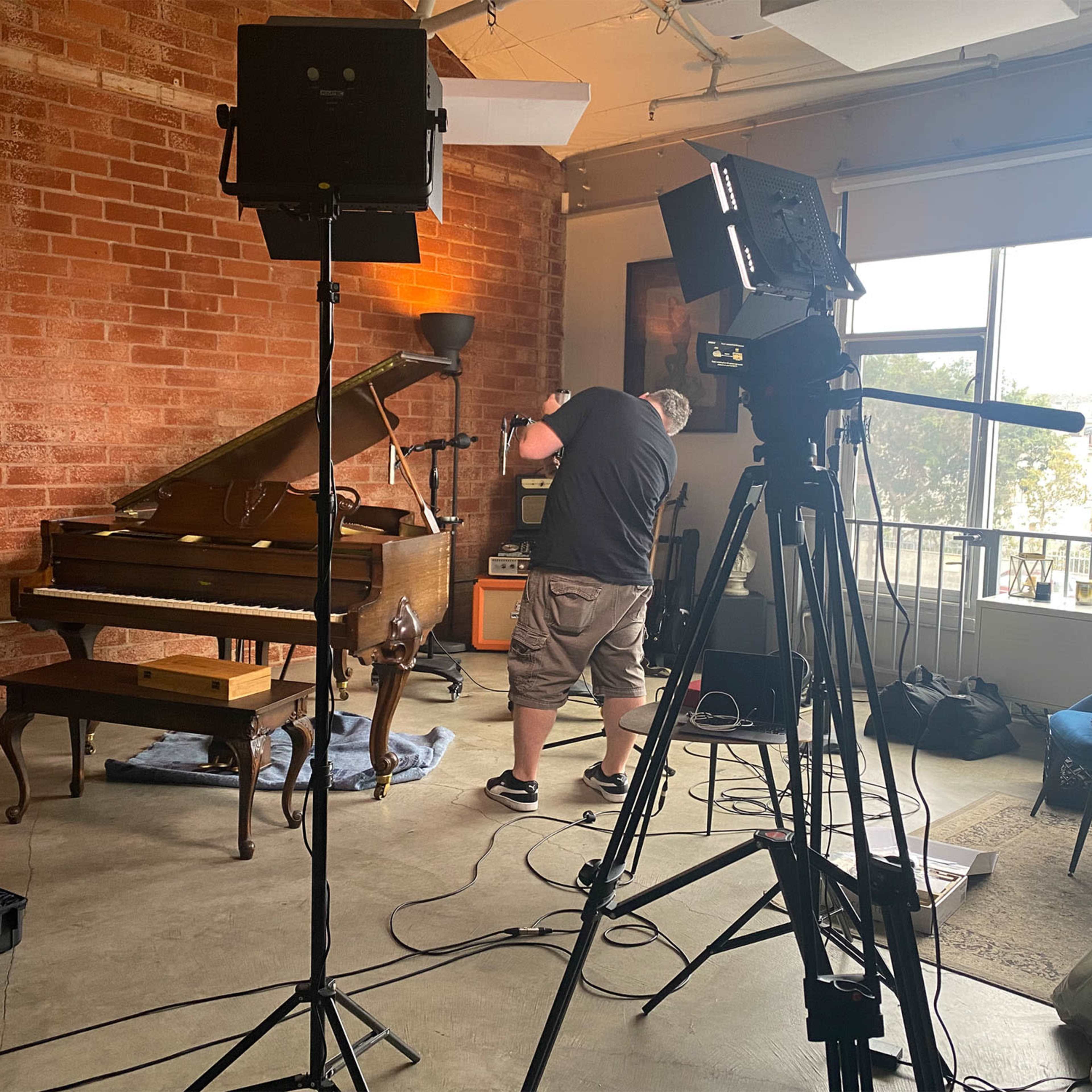 A person adjusts equipment near a vintage piano in a studio setting with exposed brick walls and soft lighting.