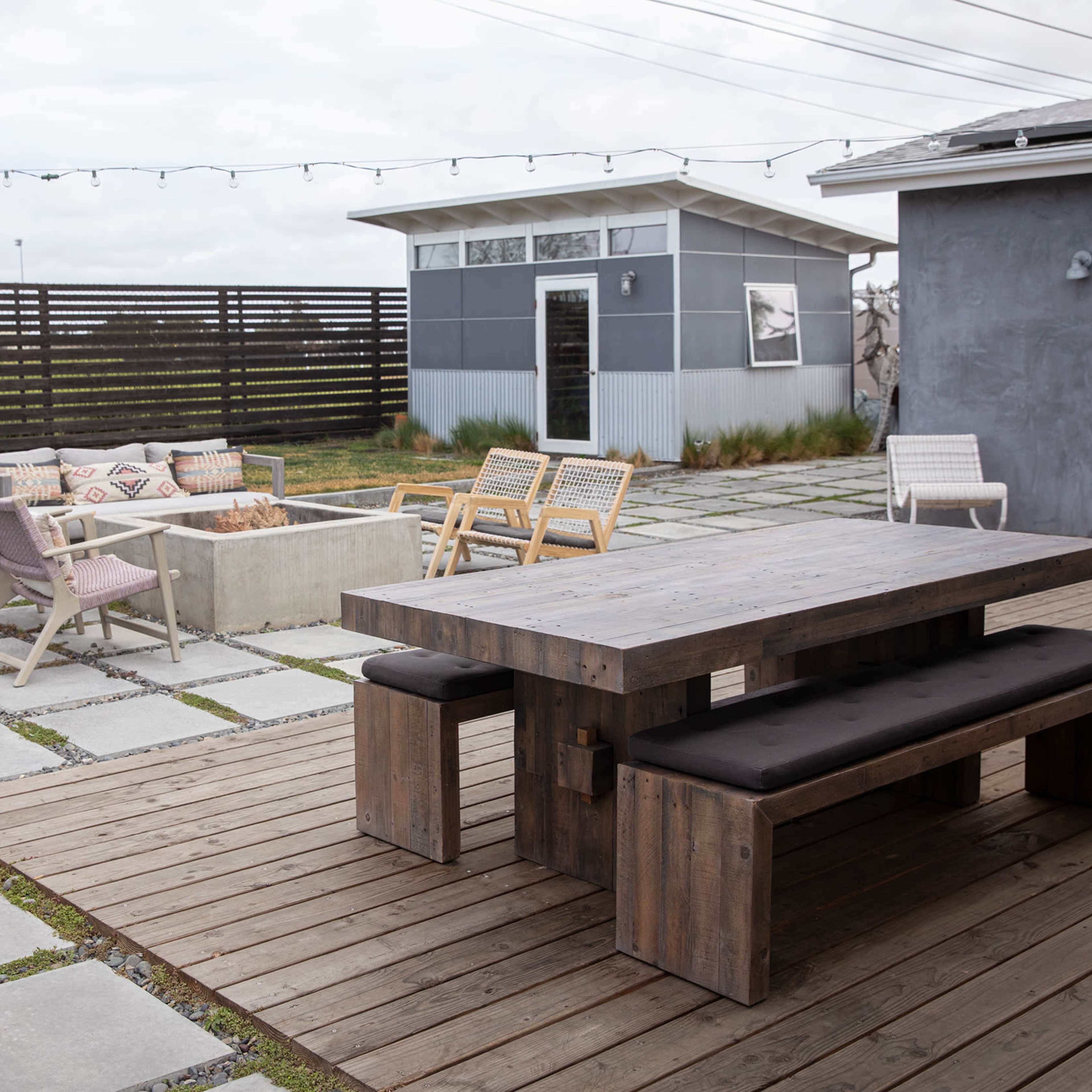 A modern outdoor patio with a large wooden table, benches, and a fire pit surrounded by various seating options, set against a gray building and a fenced area.