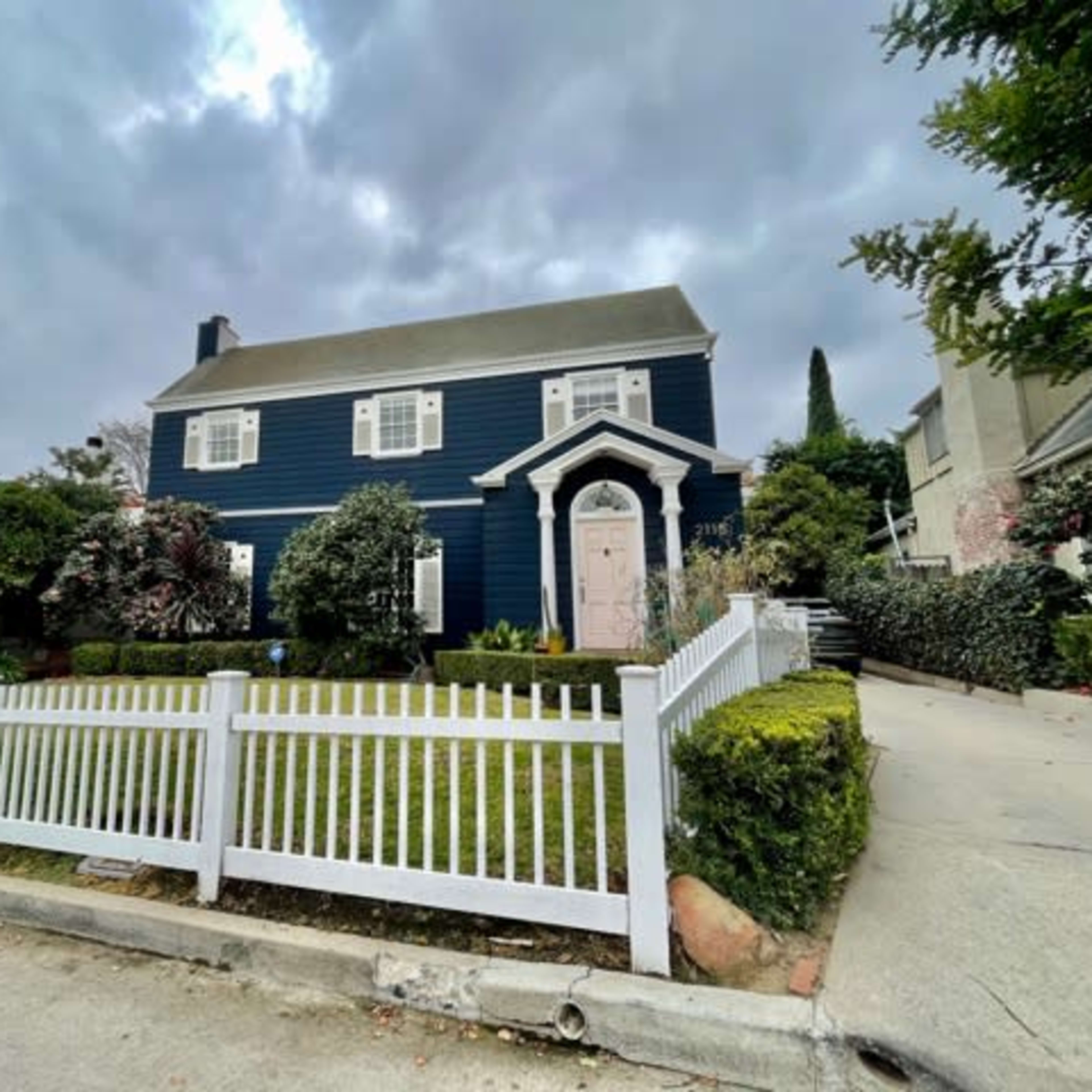 A two-story blue house with white trim and a pink door is surrounded by a white picket fence and a neatly maintained yard.