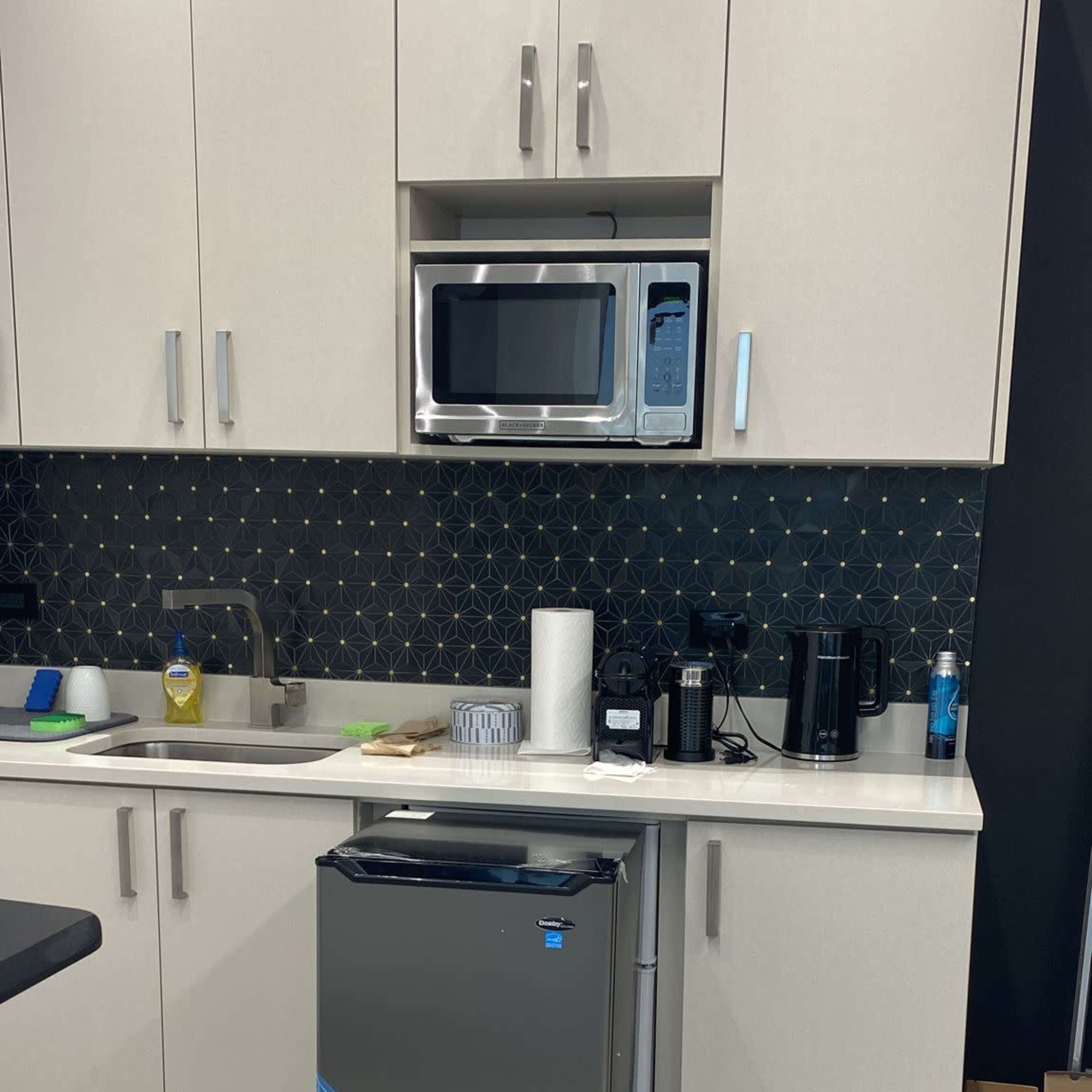 A compact silver refrigerator stands on a wooden base in a modern kitchen with light-colored cabinets and a microwave above it.