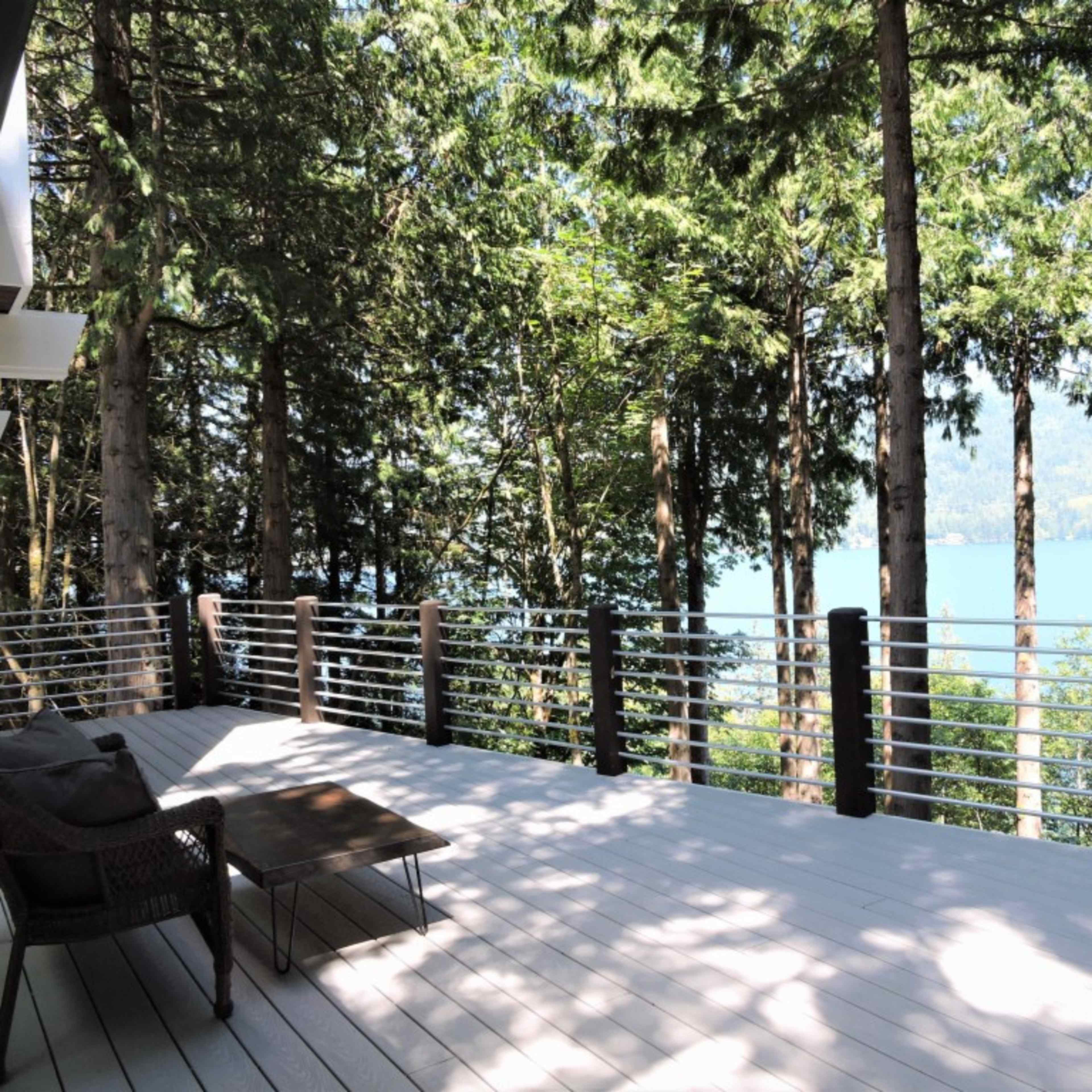 A spacious deck with a table and seating overlooks a lake, surrounded by tall trees.