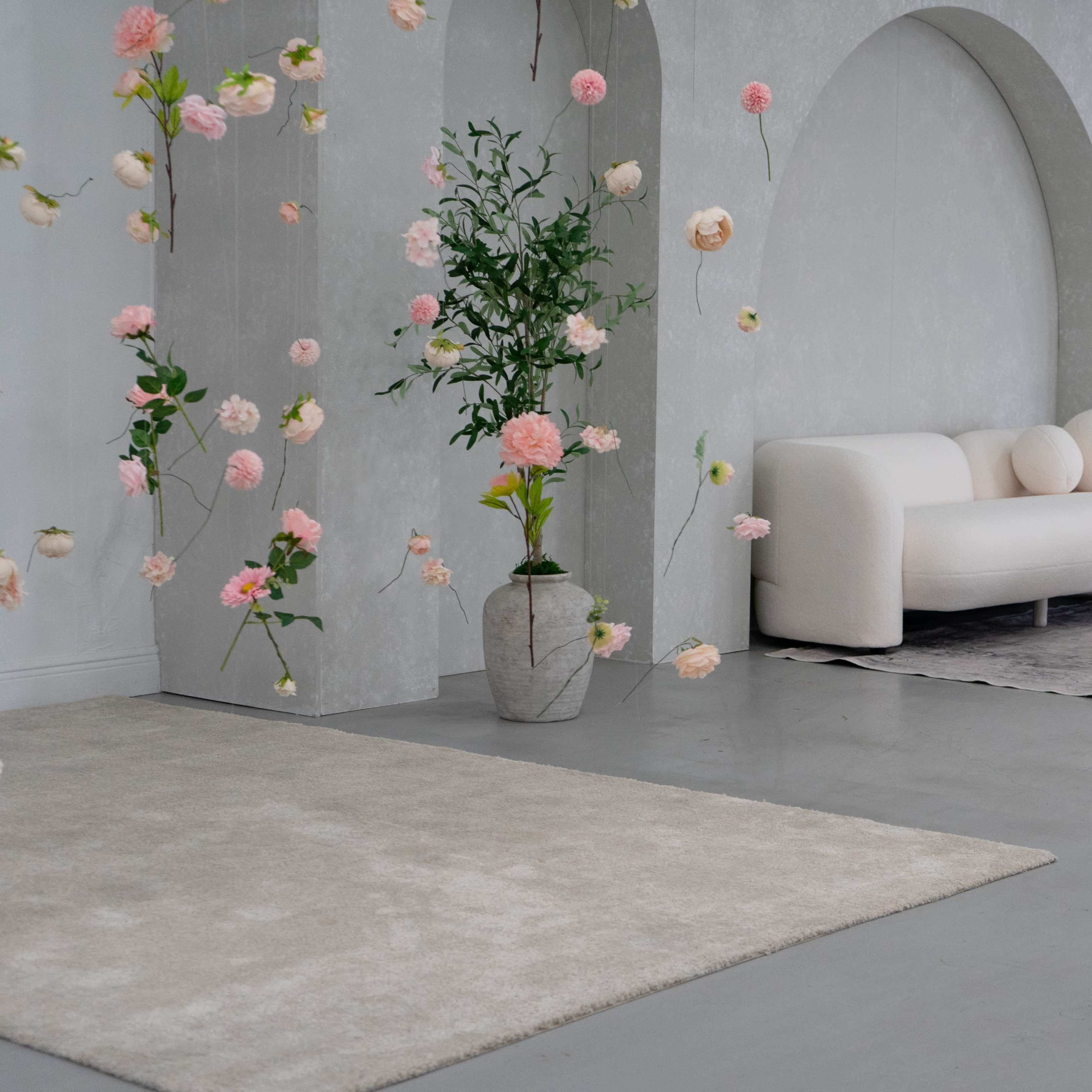 A minimalist interior with a curved white sofa, a patterned rug, and floral decorations hanging in front of a textured gray wall.