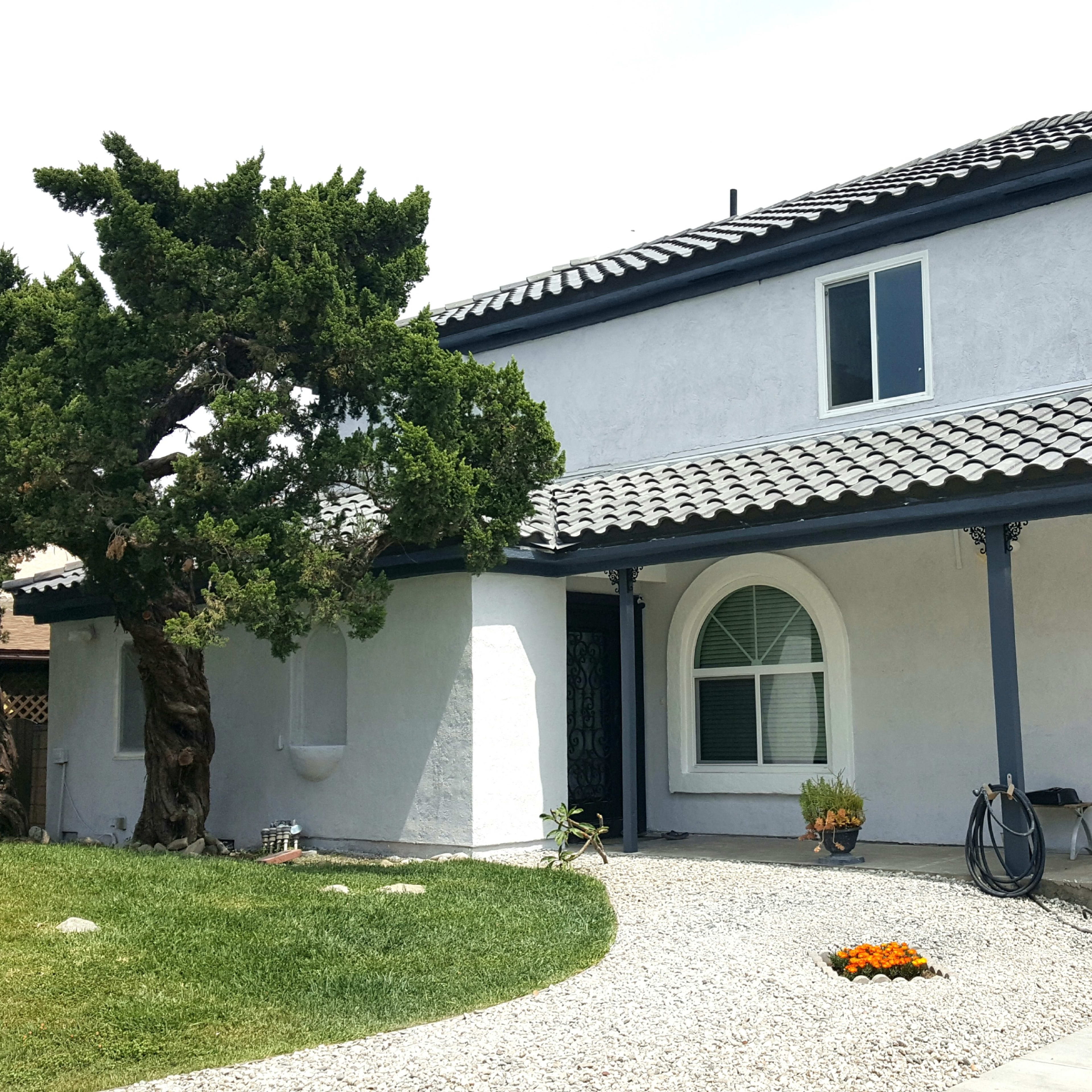 A two-story white house with a tiled roof, a front yard featuring gravel landscaping and an orange flower bed, and a large tree on the side.
