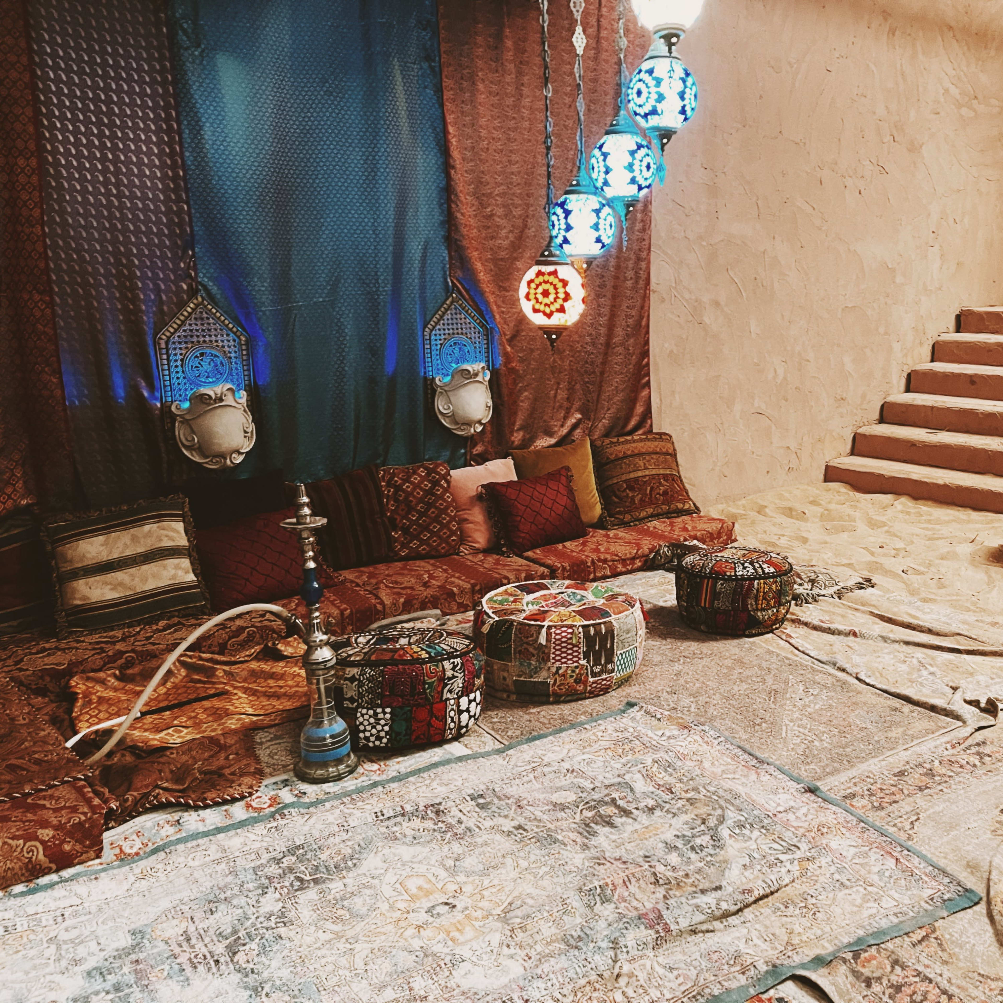 A decorated interior room with colorful cushions and rugs, ornate lamps hanging from the ceiling, and a staircase leading to a different level.