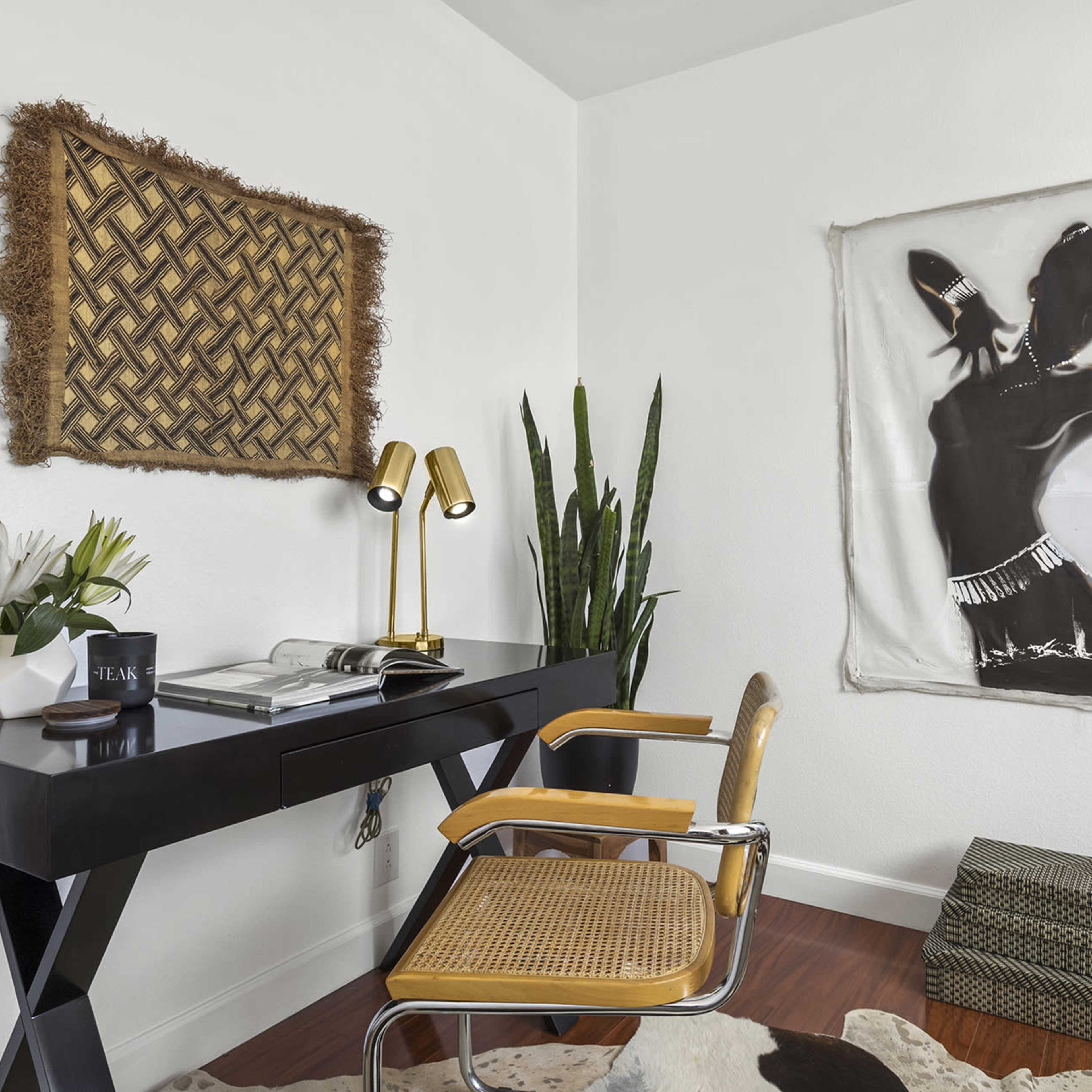 A minimalist workspace with a black desk, a chair with a woven seat, a potted plant, and a wall art piece featuring a silhouette of a figure.
