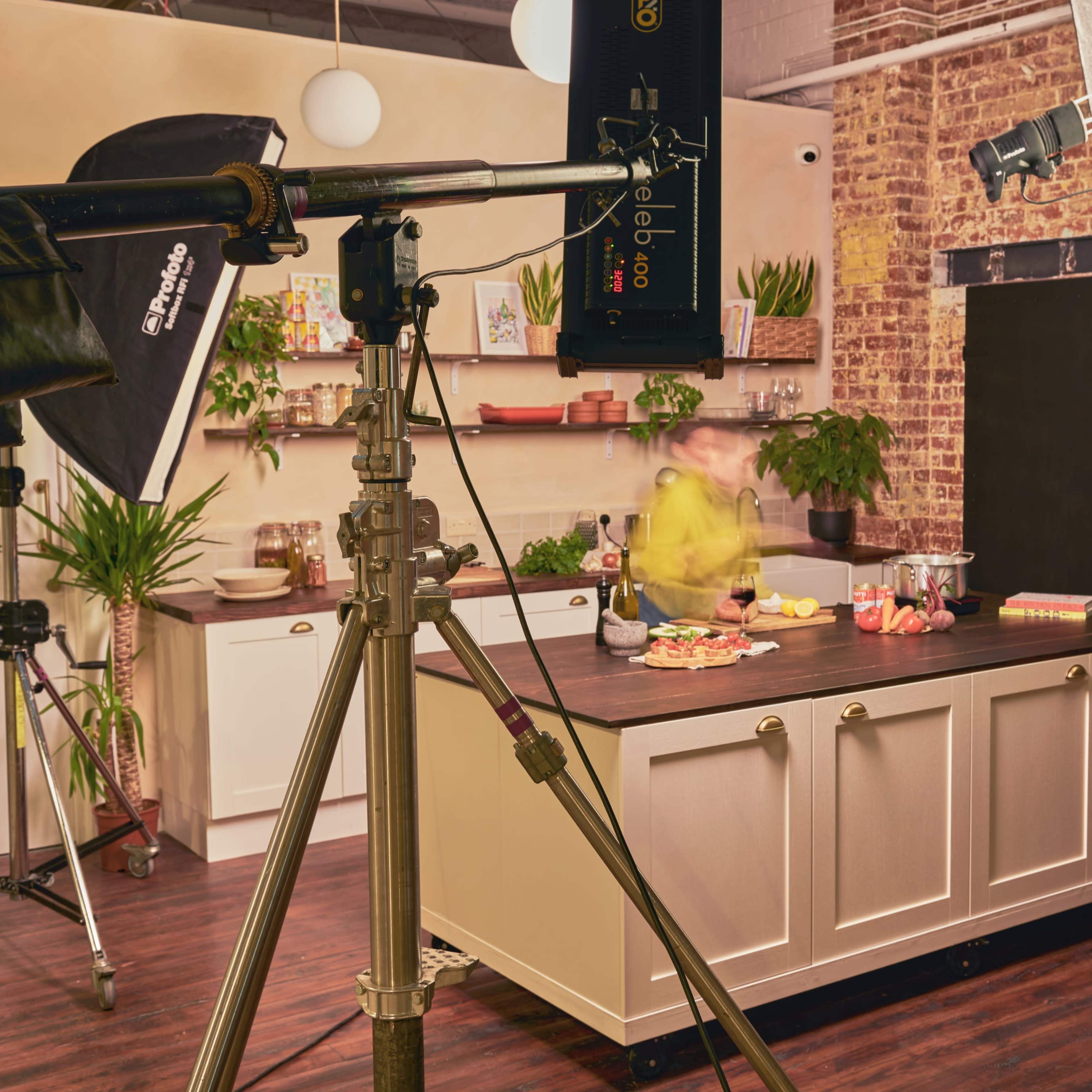 A kitchen photography setup features a well-lit countertop with food and drinks, surrounded by studio lighting and camera equipment.