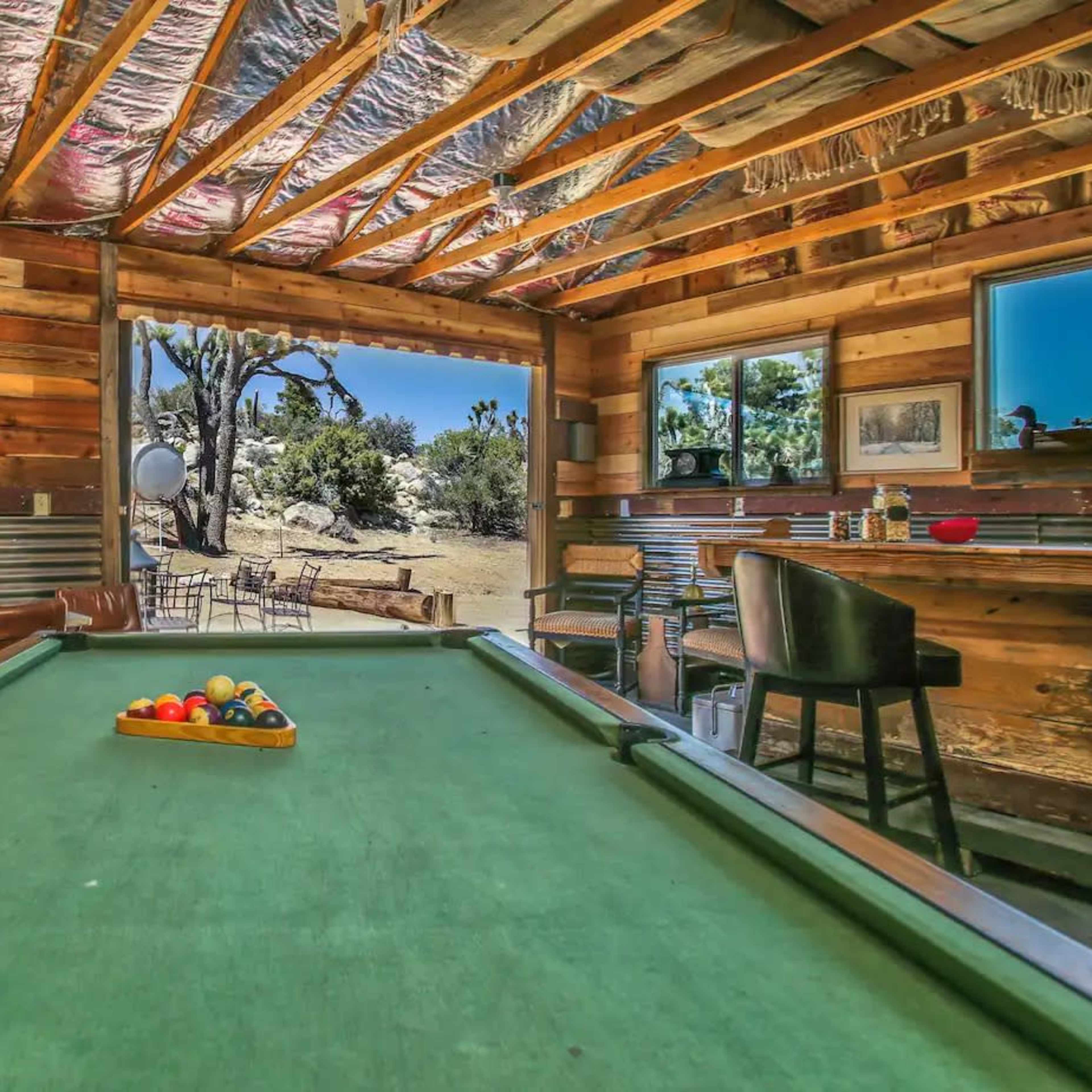 A rustic game room with a green pool table in the foreground and large windows revealing a desert landscape outside.