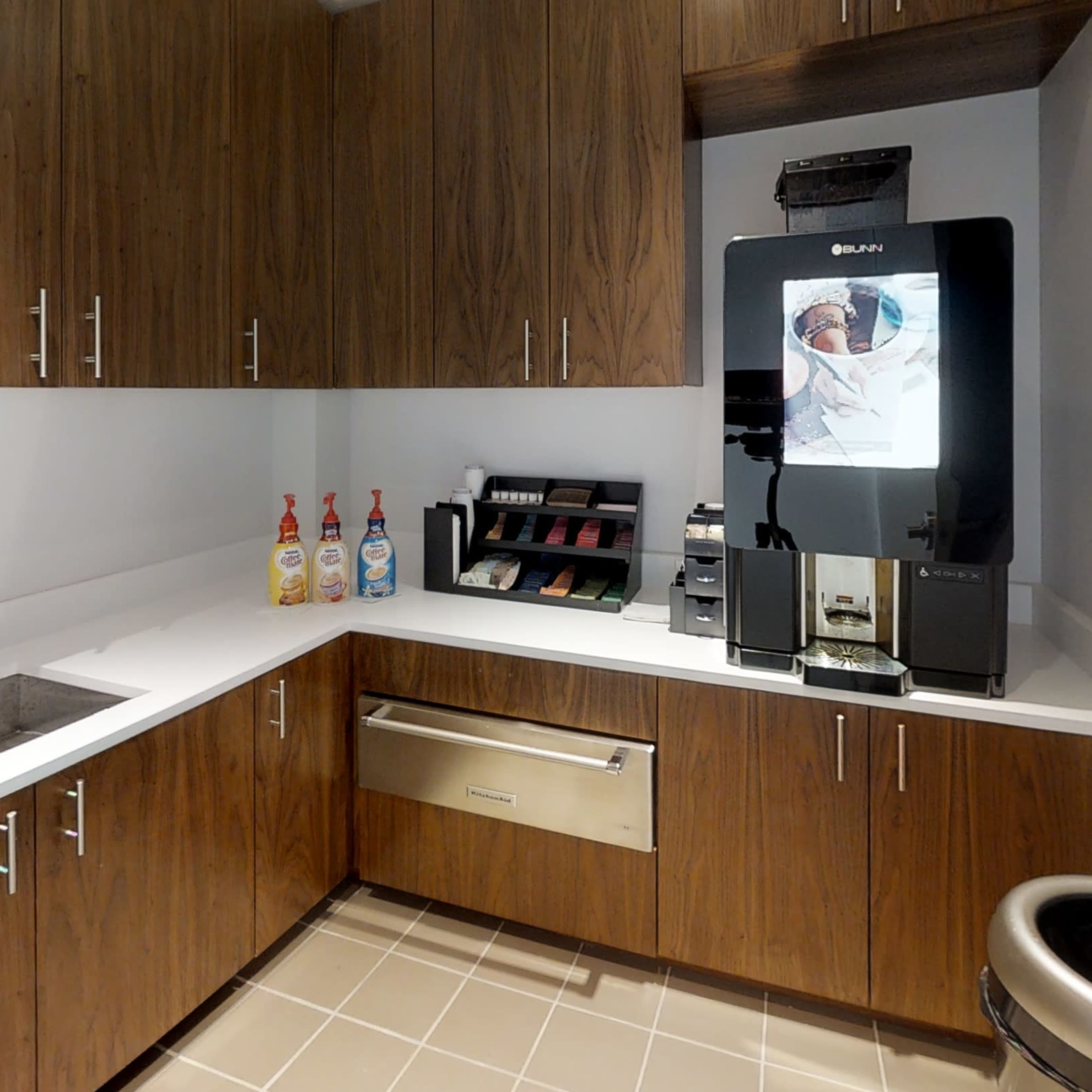 A kitchen space with wooden cabinets, a sink, a countertop, a beverage dispenser, and a trash can.