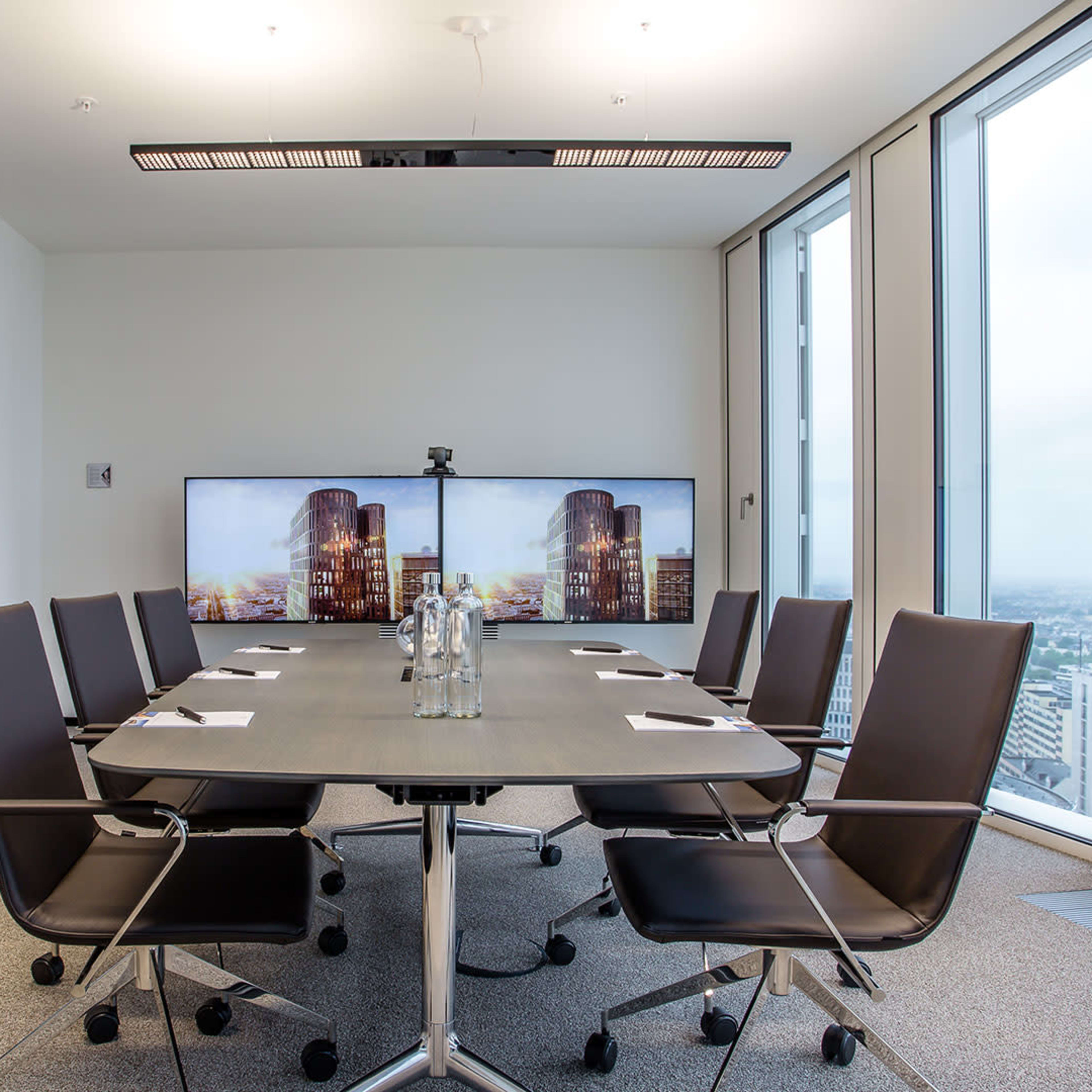 A modern conference room with a rectangular table, black chairs, two large screens displaying cityscapes, and a large plant in a corner.