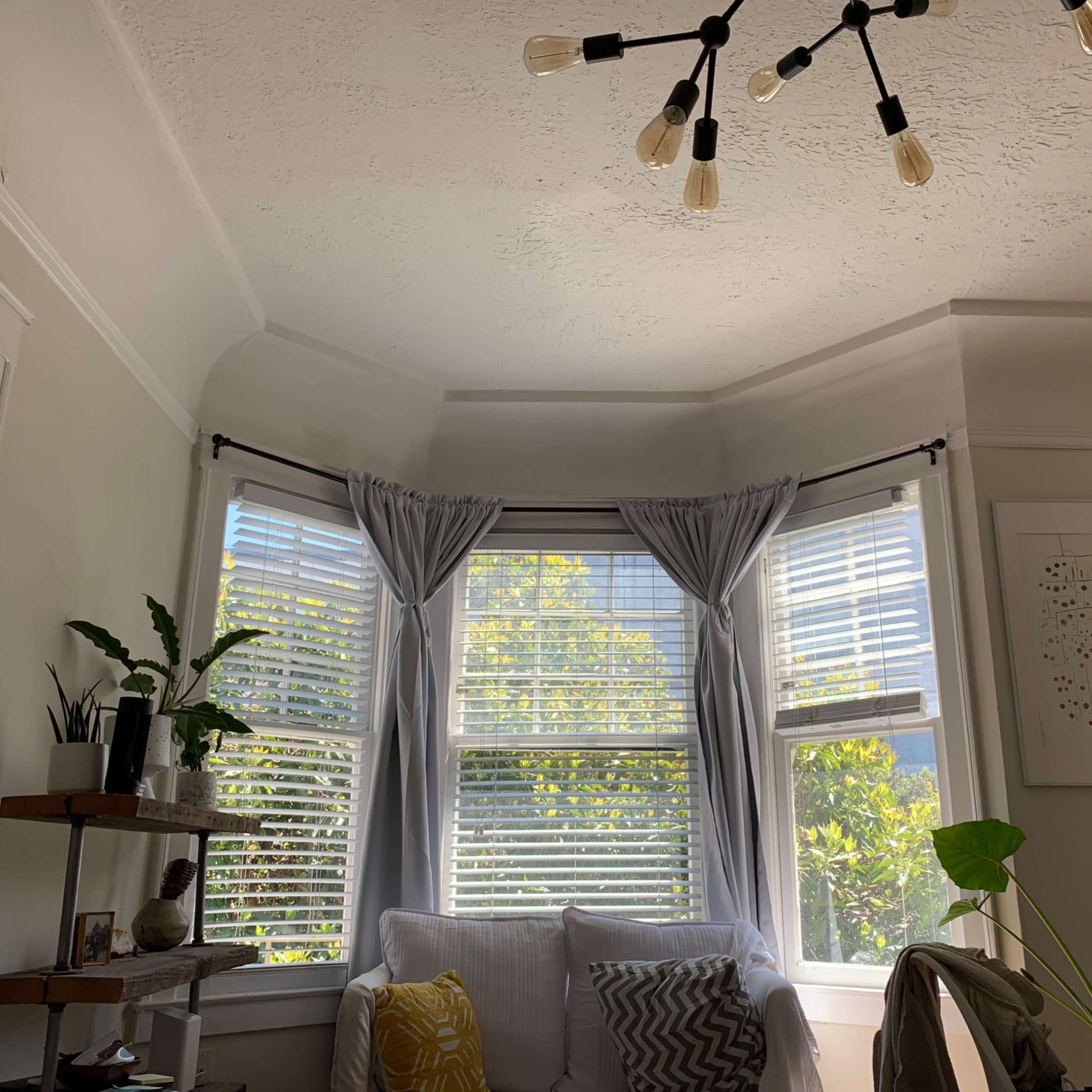 A cozy living room corner with a bay window adorned with light curtains, a small plant on a shelf, and a white sofa.