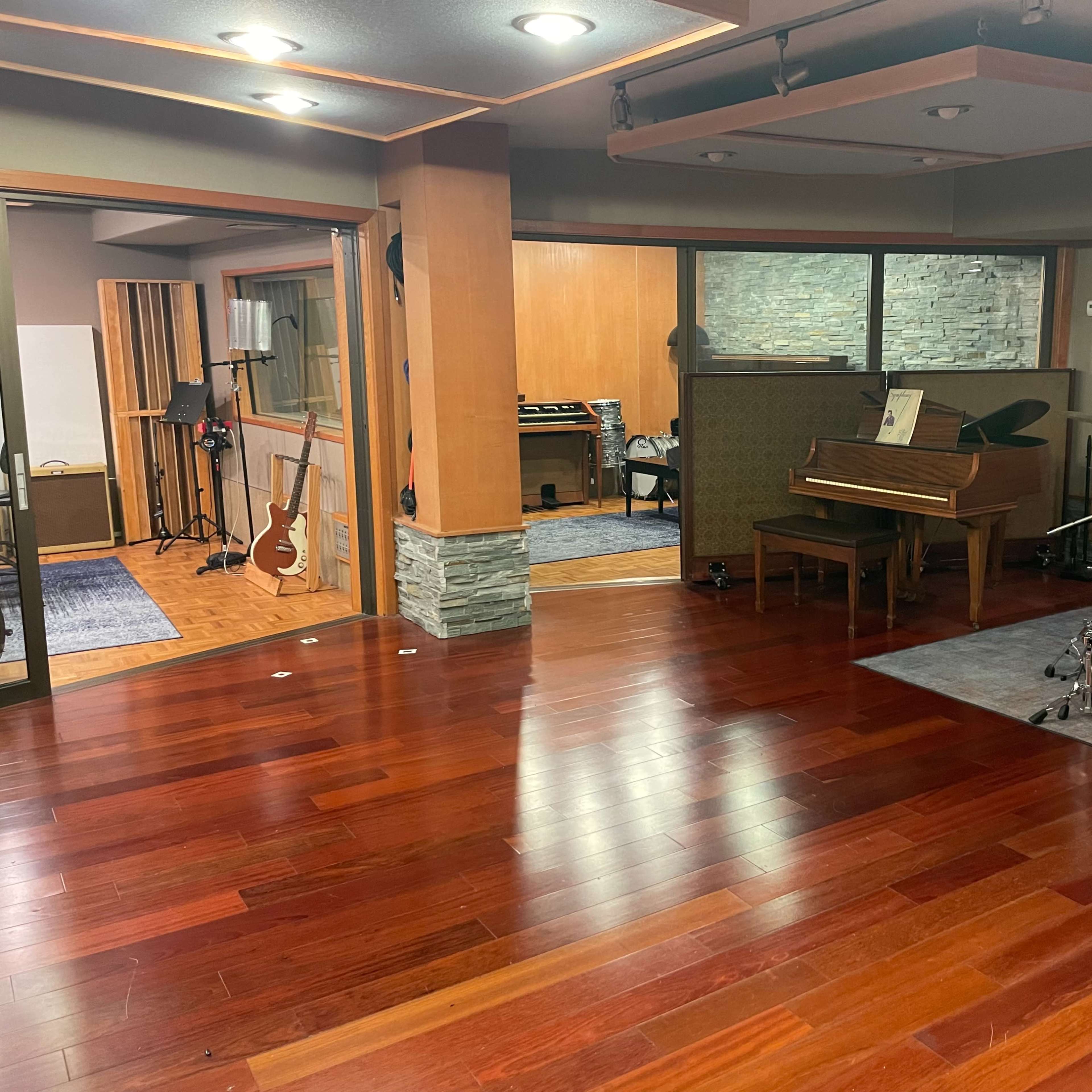 A music studio features a wooden floor, a grand piano, a drum set, and various recording equipment visible in the background.
