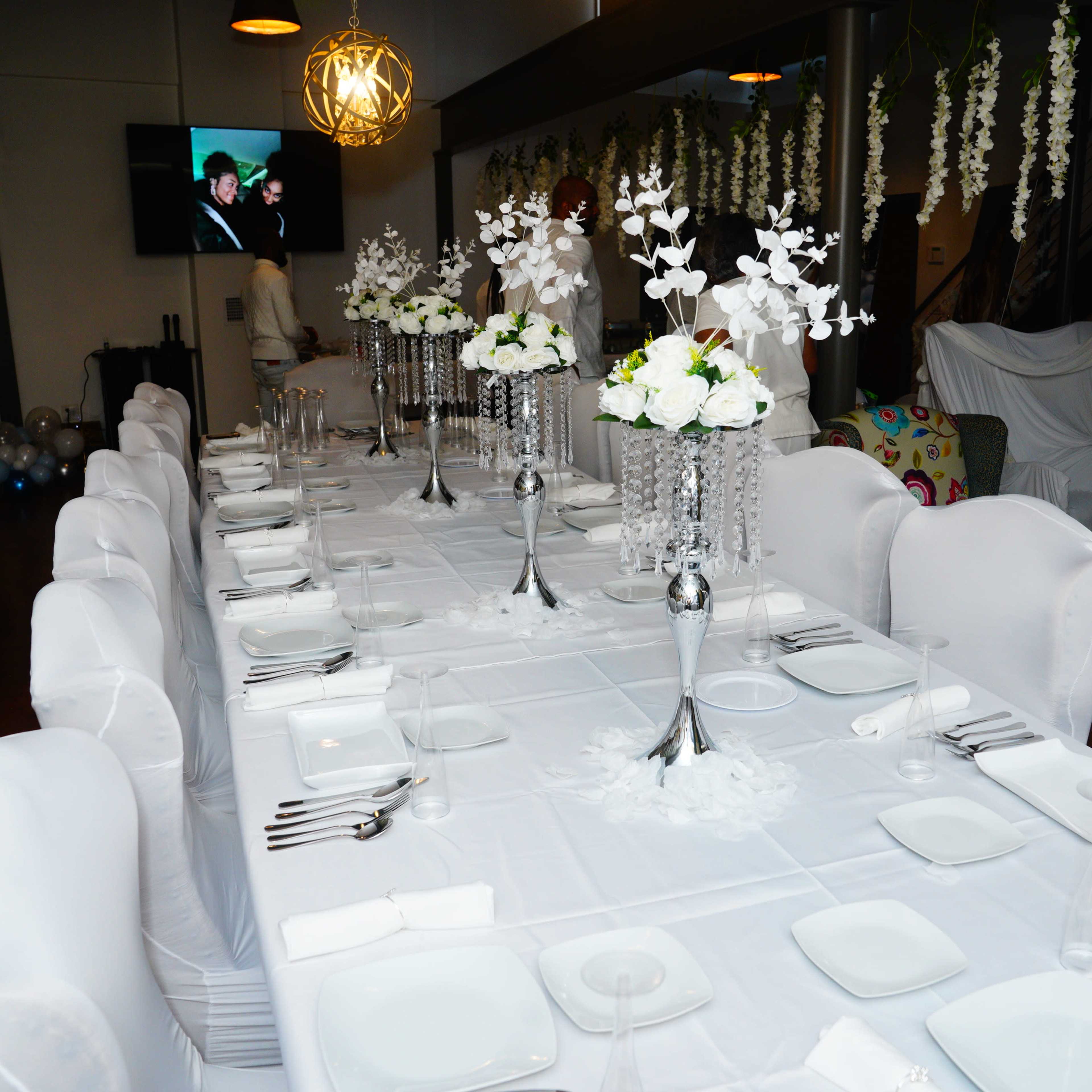 A long, elegantly set dining table with white decorations, featuring floral centerpieces and a television playing in the background.