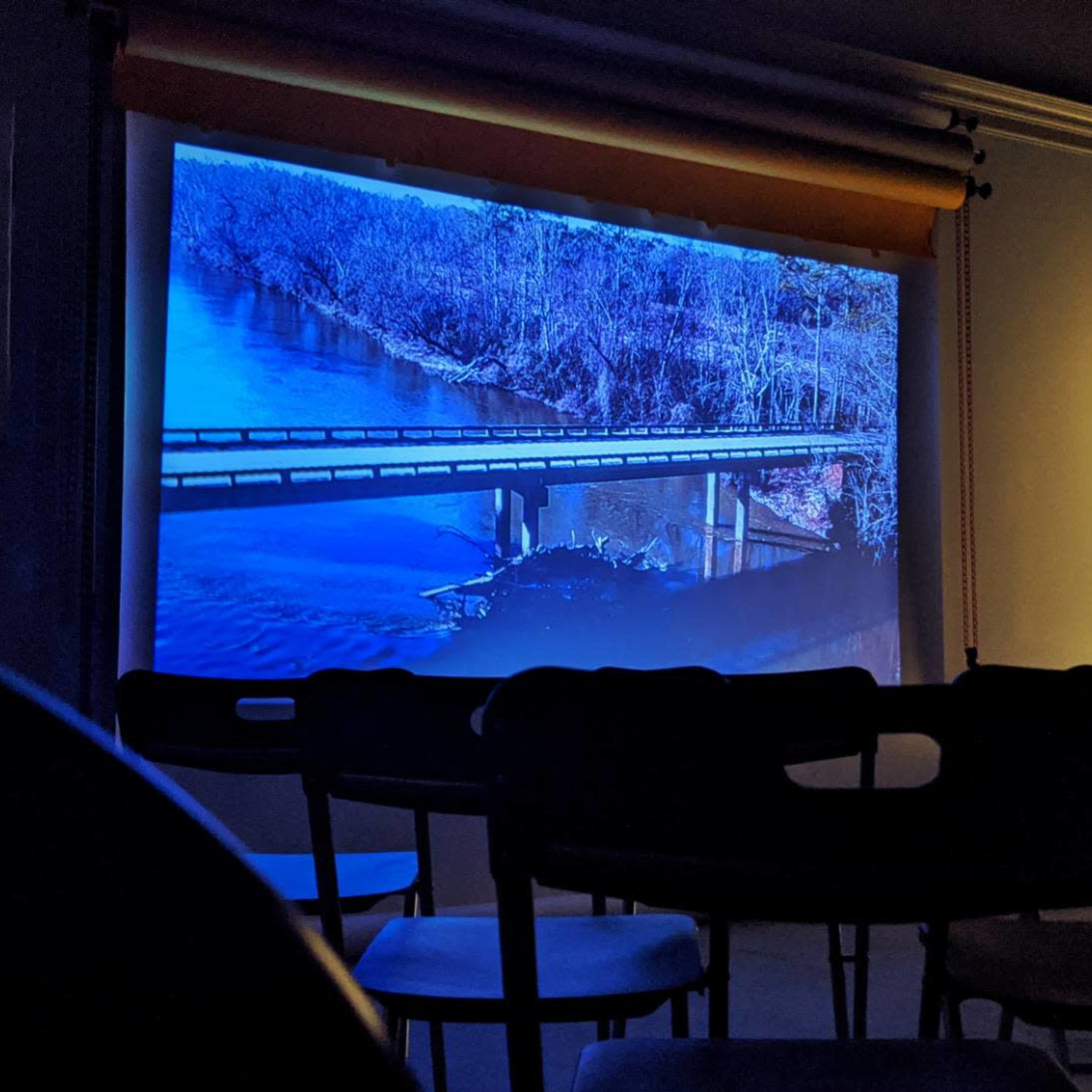 A projection of a river and bridge scene is displayed on a screen in a dimly lit room with empty chairs arranged in front of it.
