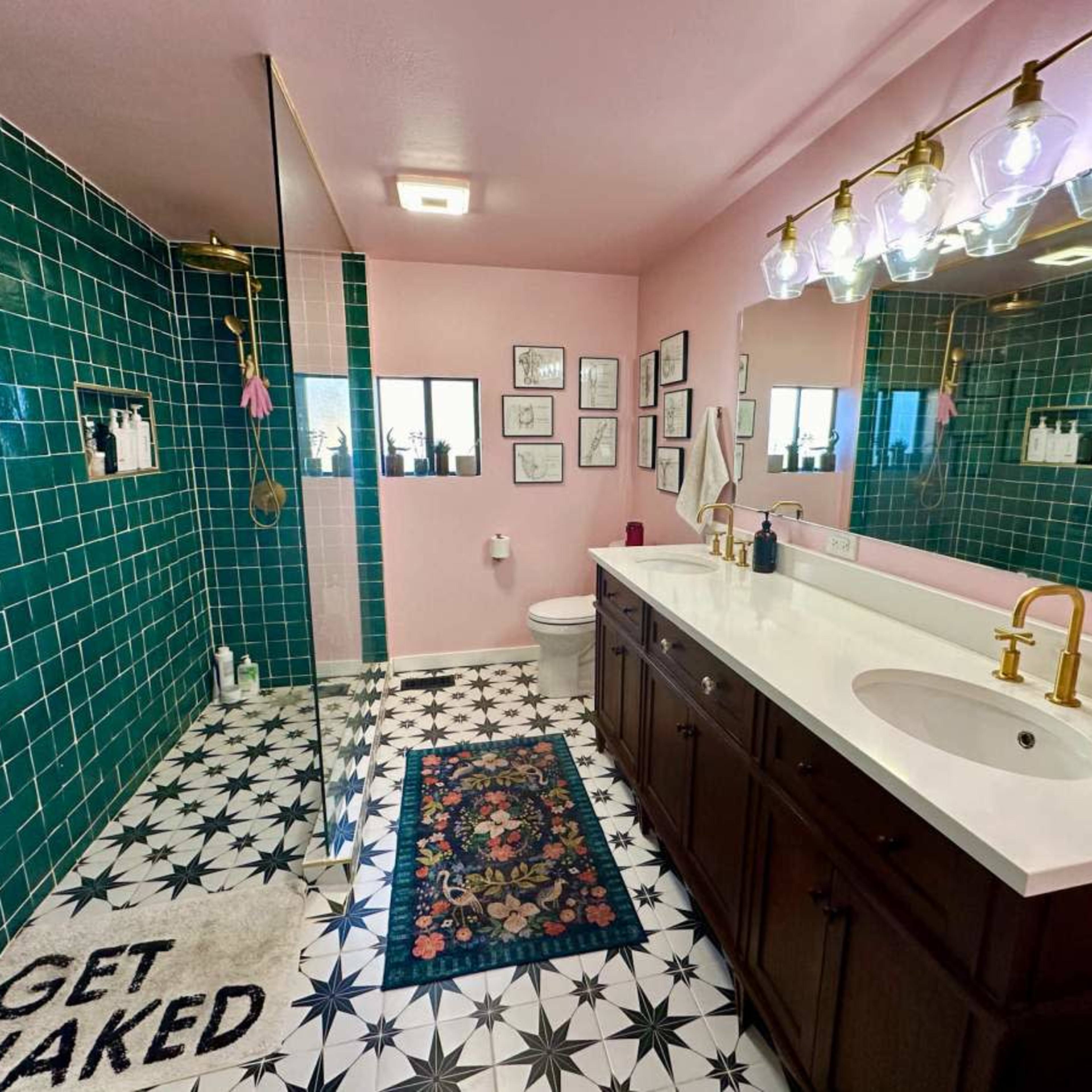 The bathroom features a spacious layout with green tiled walls, a glass shower, a double vanity with gold fixtures, and a patterned floor.