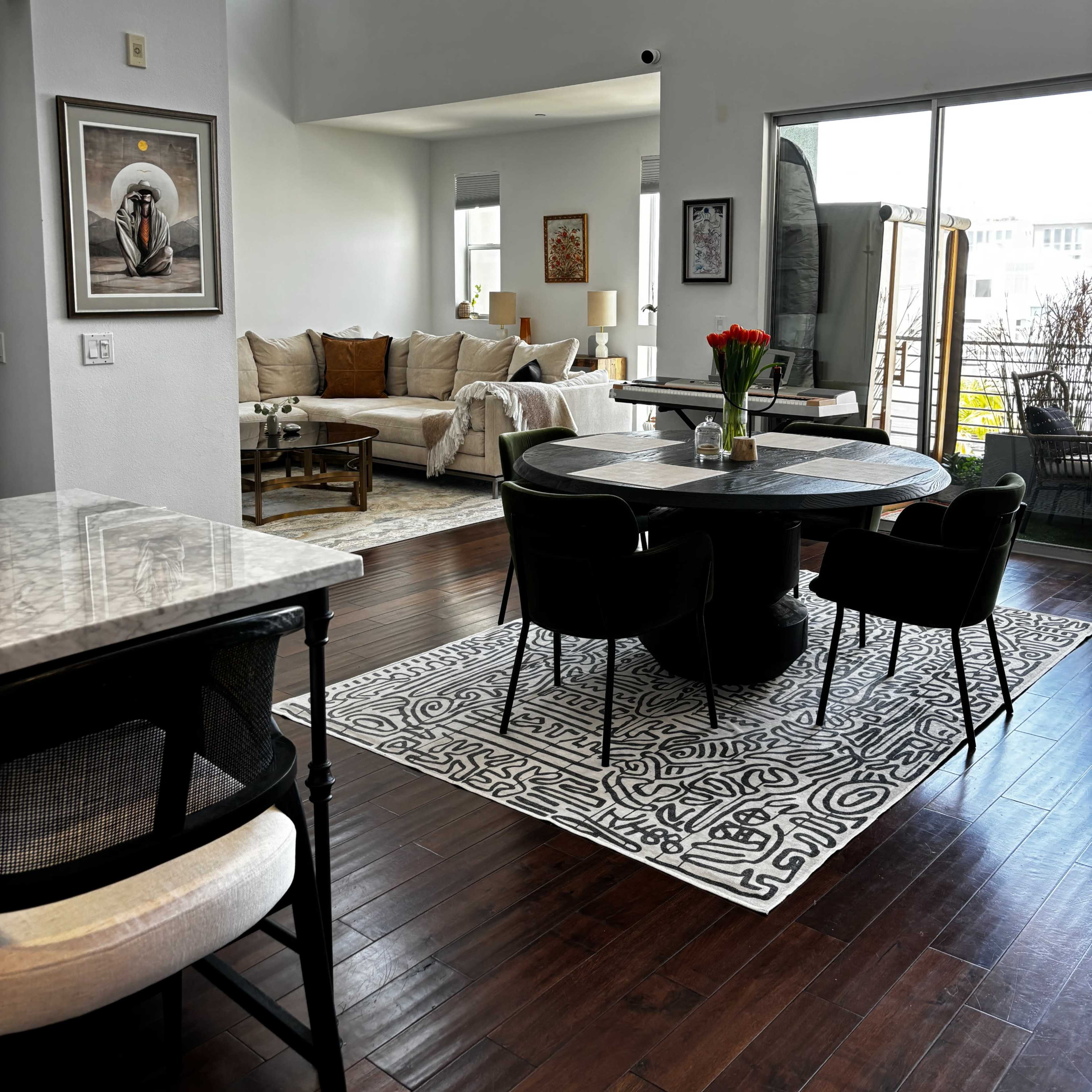 A spacious living area features a dining table with black chairs, a gray rug, and a comfortable seating arrangement under large windows.