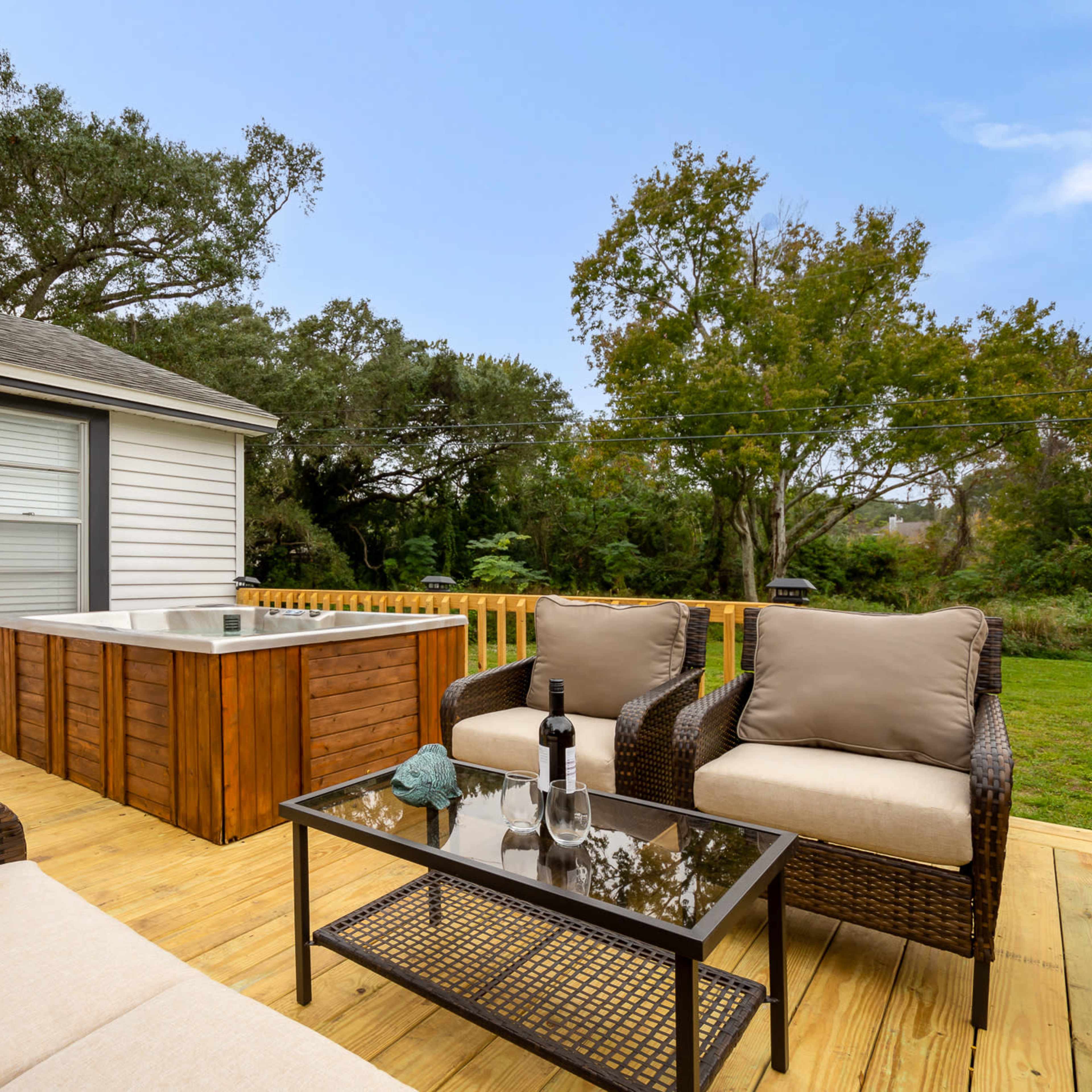 A deck features a seating area with two upholstered chairs and a glass-top table, alongside a hot tub framed by greenery.