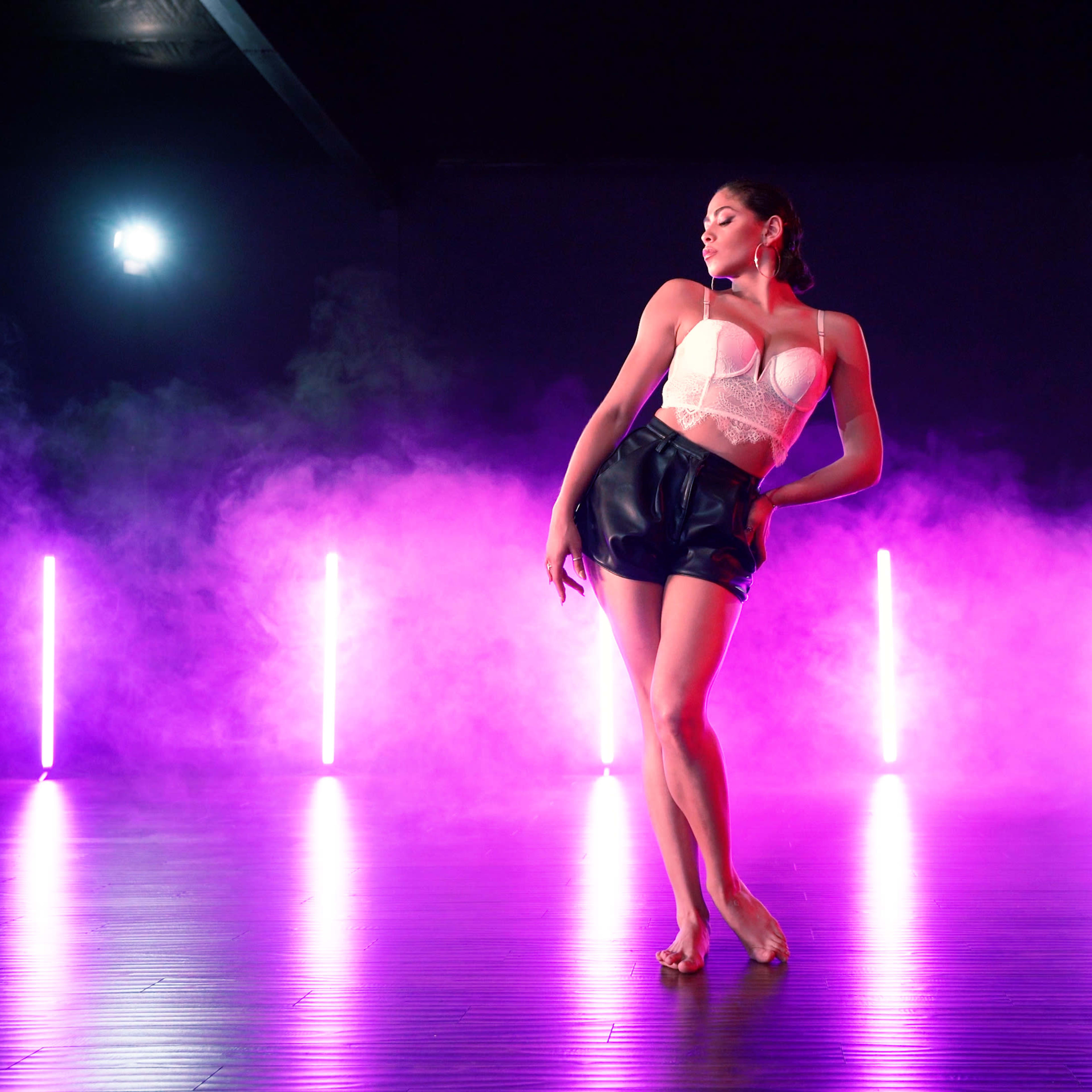 A dancer poses in a dimly lit studio surrounded by pink and purple smoke, with bright lights illuminating the scene.