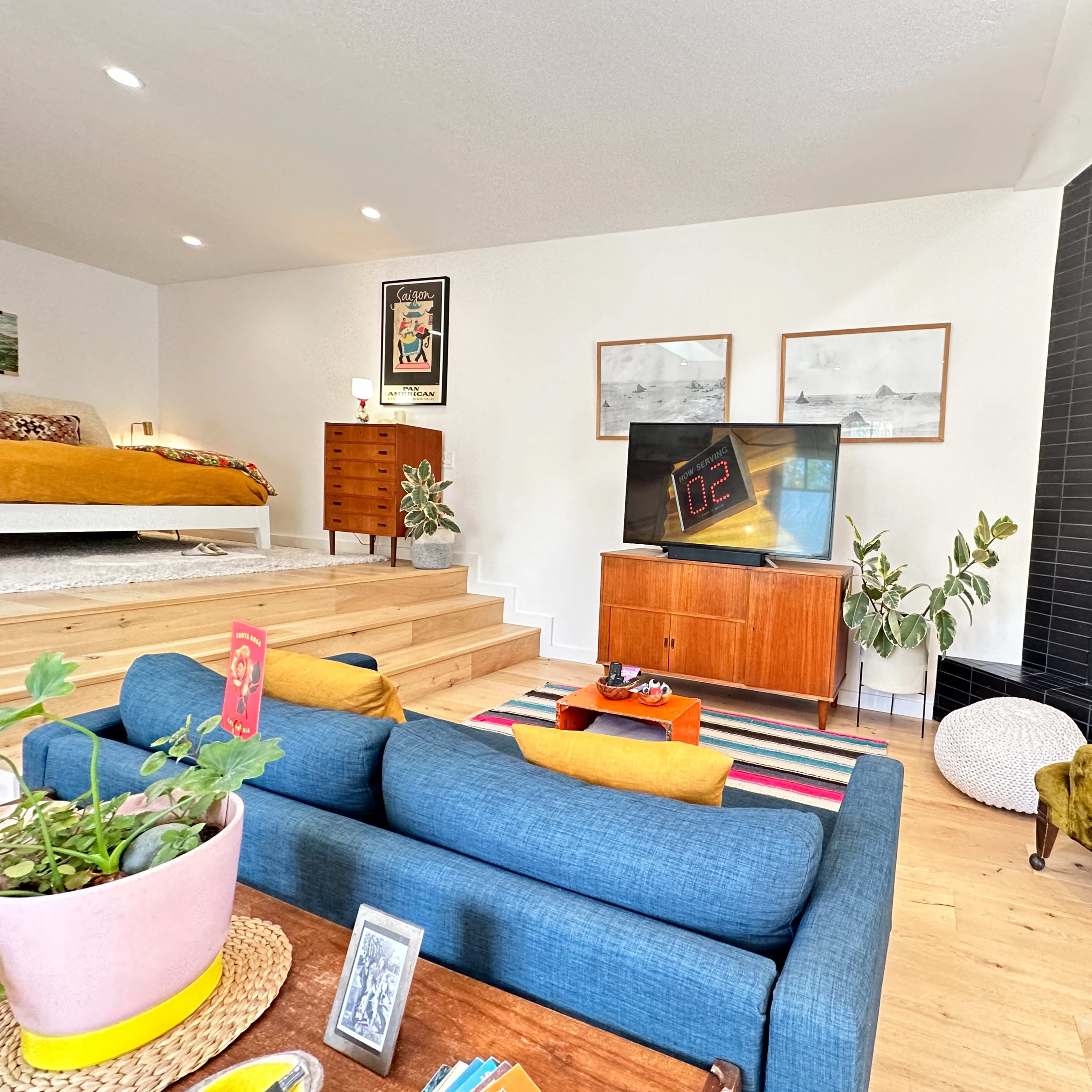 A modern living room features a blue sofa, a television on a wooden cabinet, a yellow armchair, and a staircase leading to a bedroom area.