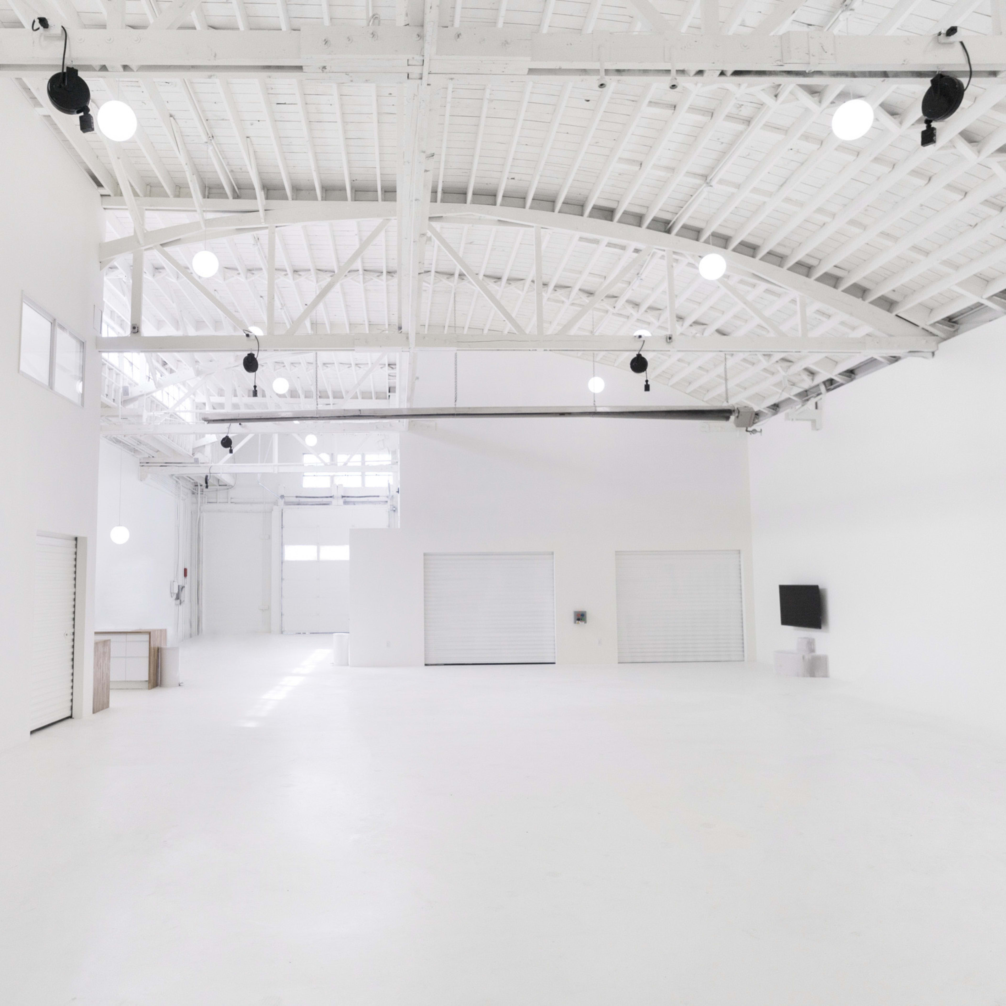 A spacious, well-lit interior with white walls, a high ceiling, and minimal furnishings, featuring large windows and double doors at one end.