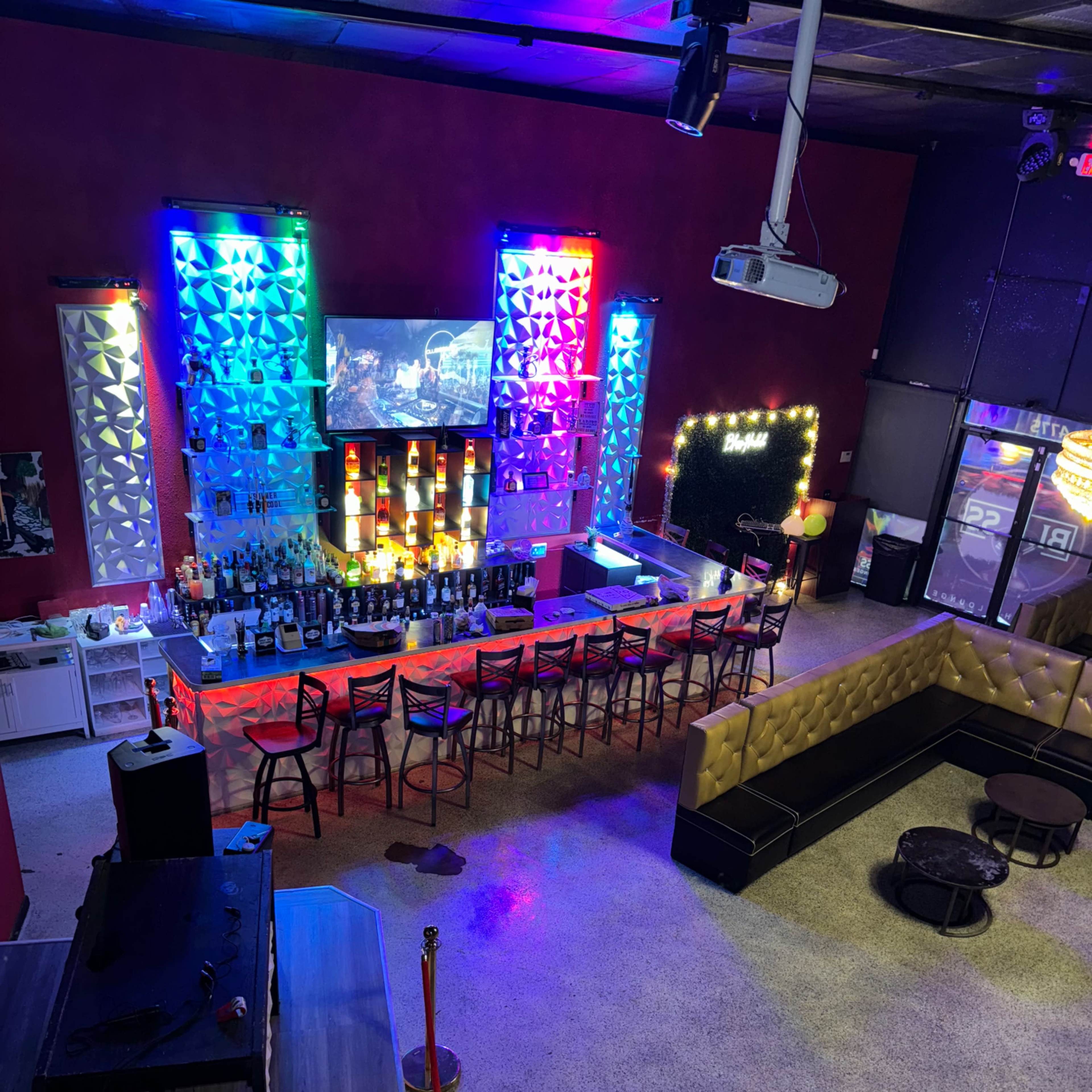 A brightly lit bar area with colorful lights, a linear bar counter lined with various bottles, and a seating section featuring black couches and gold accents.