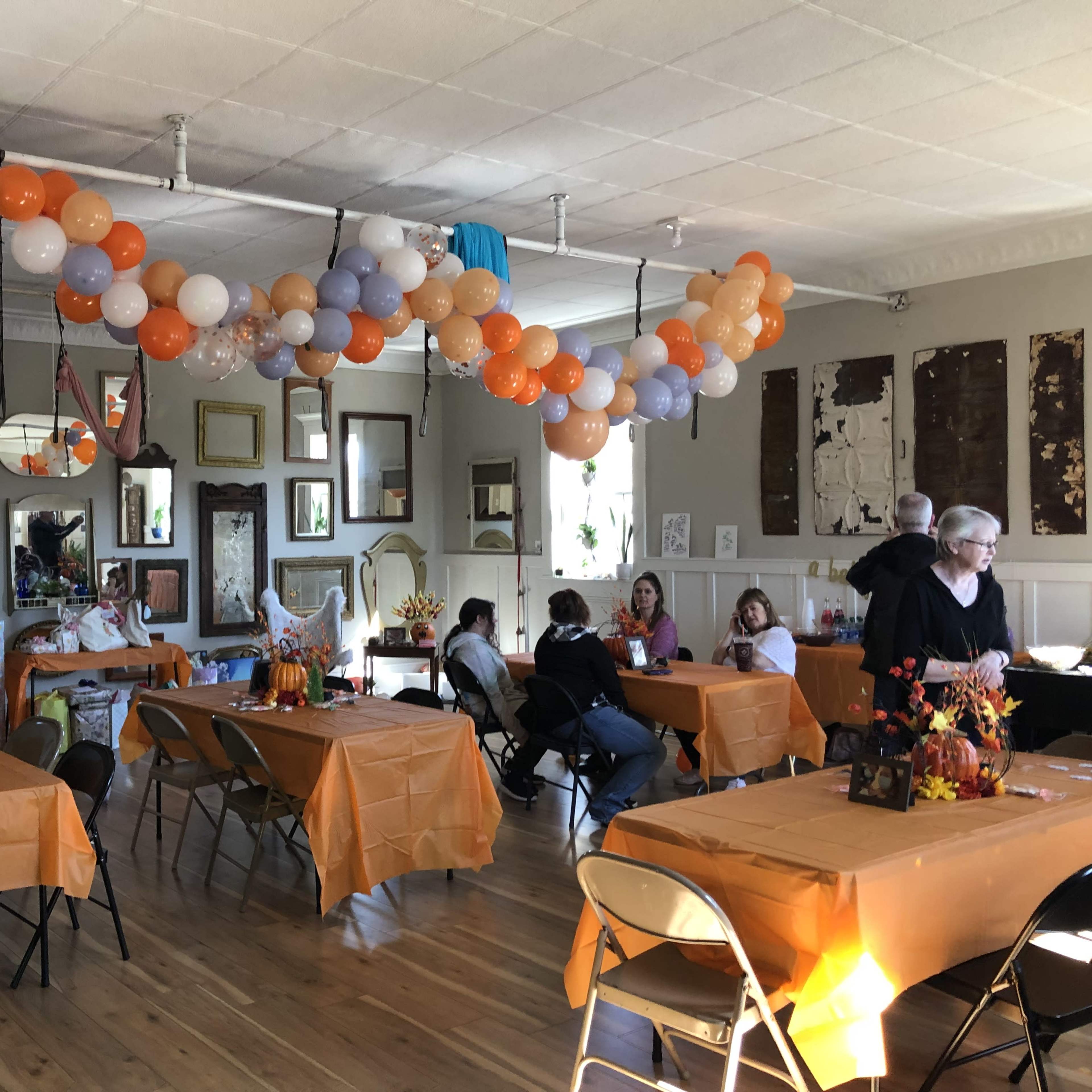 A decorated room with orange tablecloths, a balloon arch overhead, and several tables set up for a gathering.