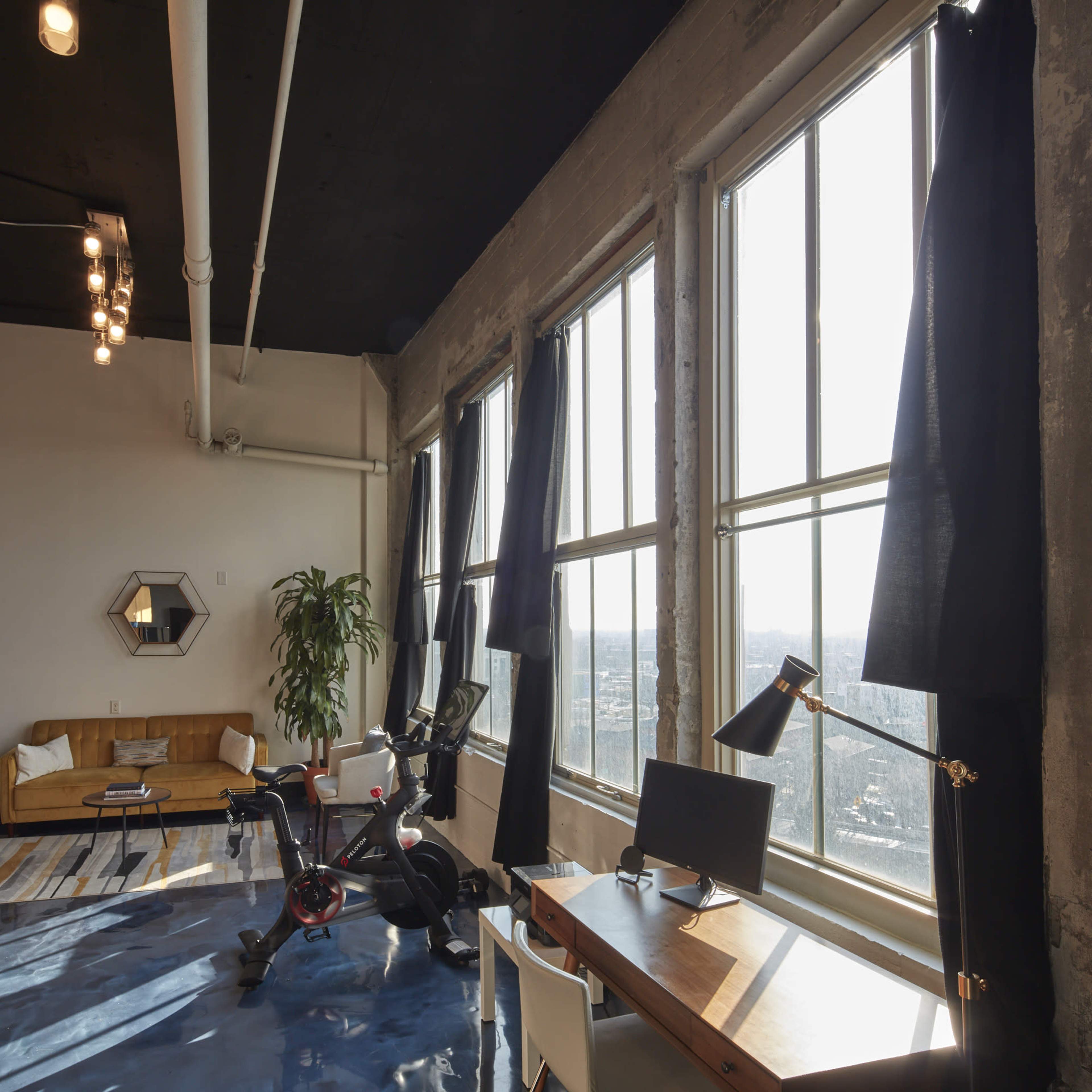 A spacious industrial-style room features large windows, a yellow couch, a bike, and a desk with a computer.