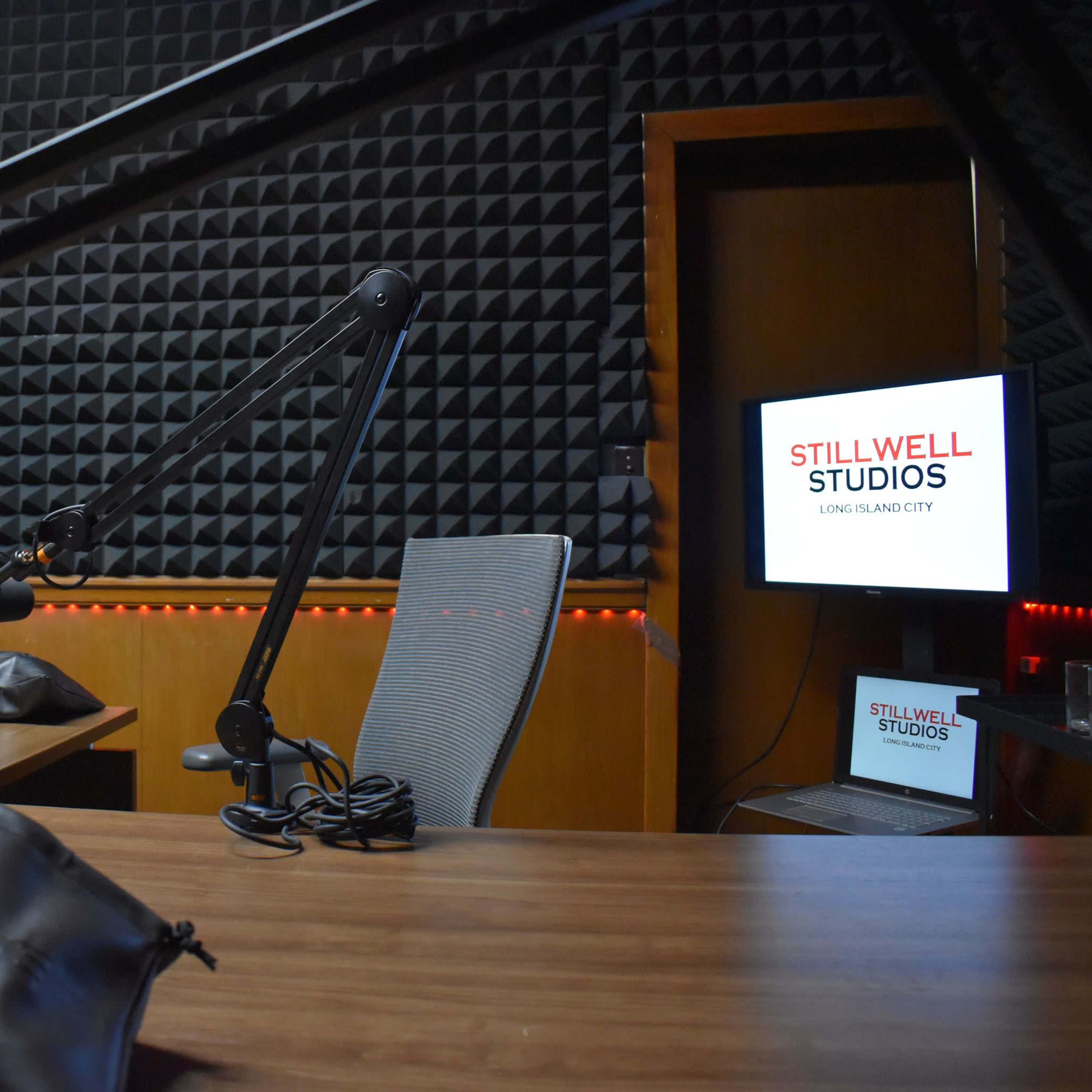 A recording studio with microphones on adjustable arms, a wooden table, and a screen displaying "STILLWELL STUDIOS" in a dimly lit room with soundproof walls.