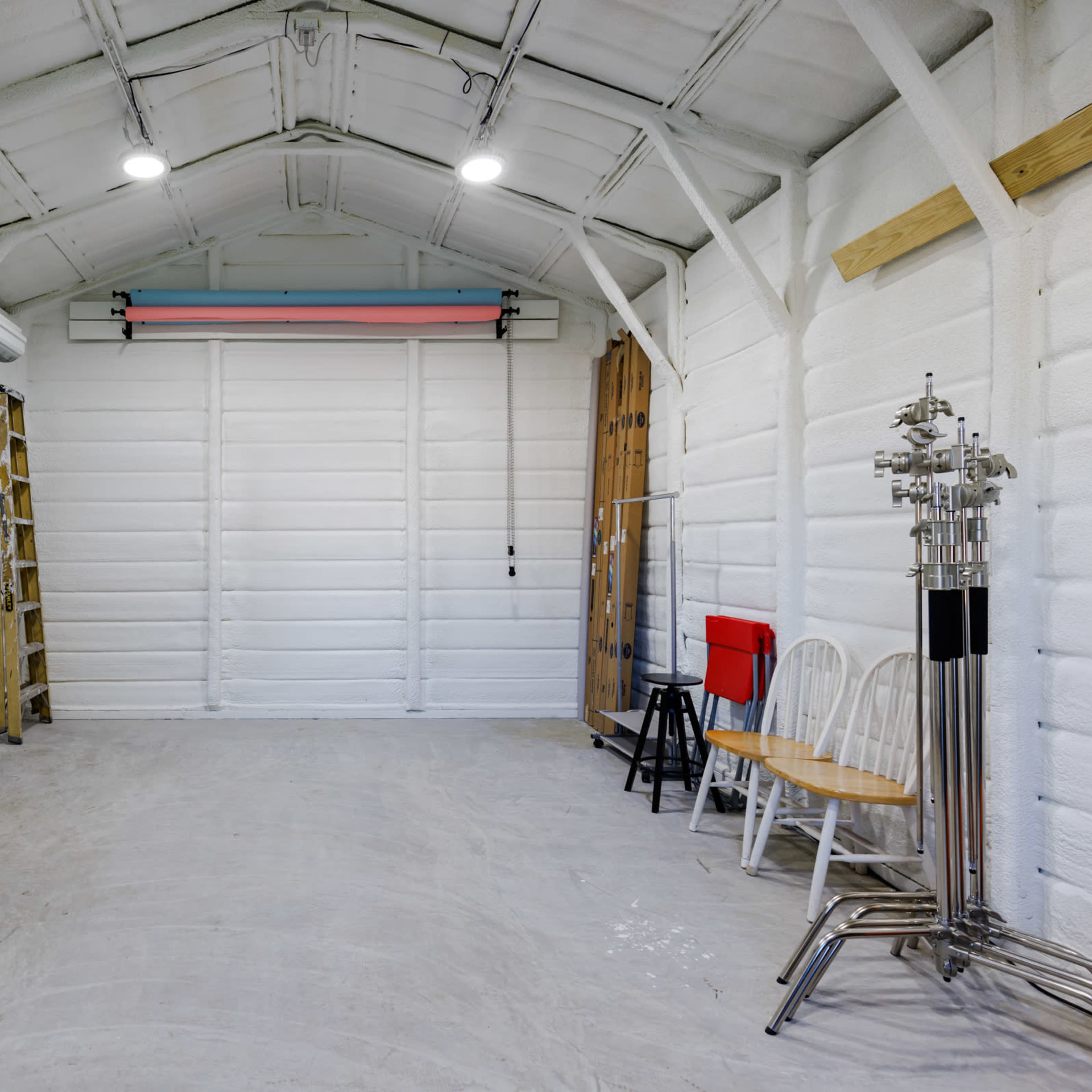 A spacious, empty garage with a roll-up door, several chairs, and ladders against the white-paneled walls.