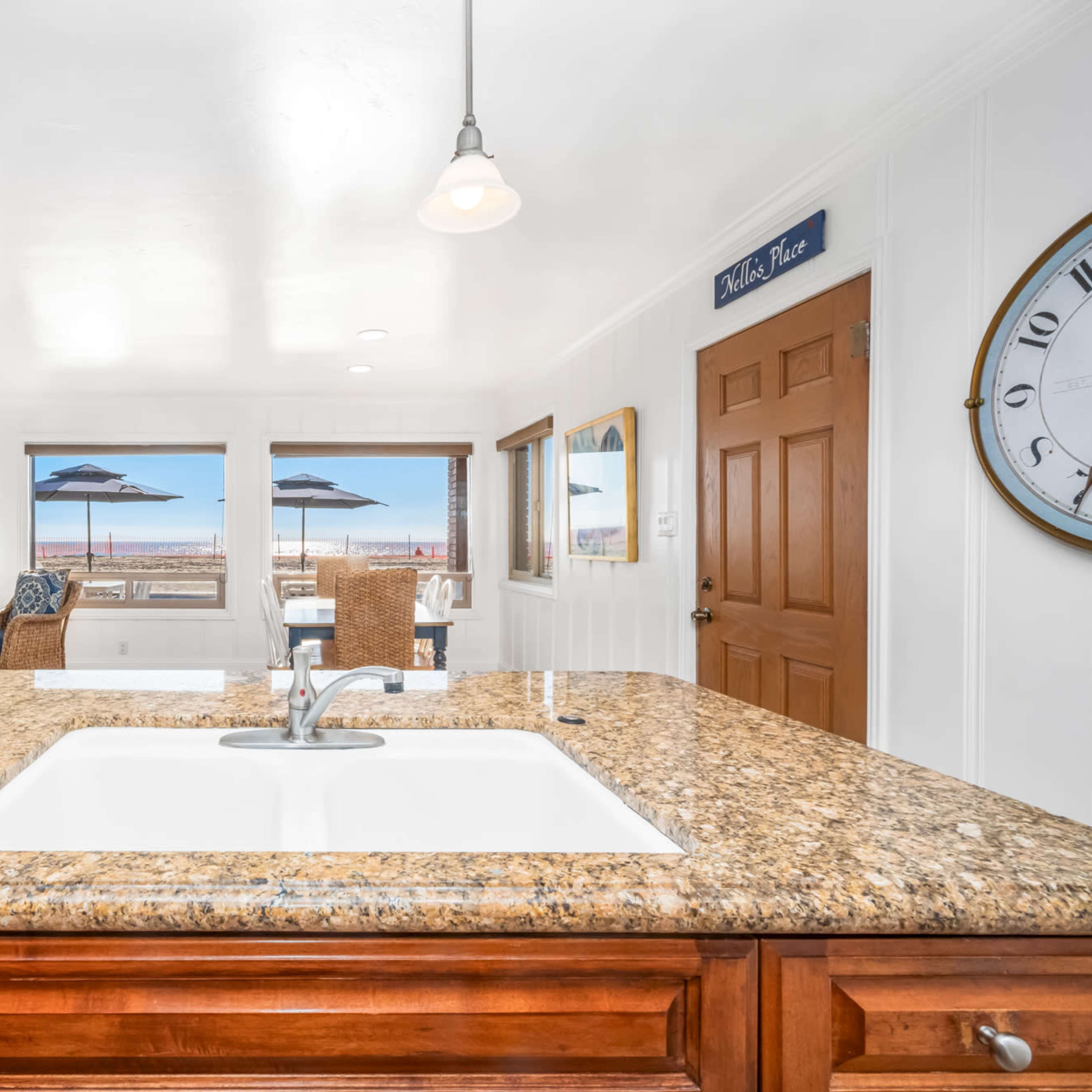 A kitchen with a granite countertop overlooks a living area with seating and a view of the beach through large windows.