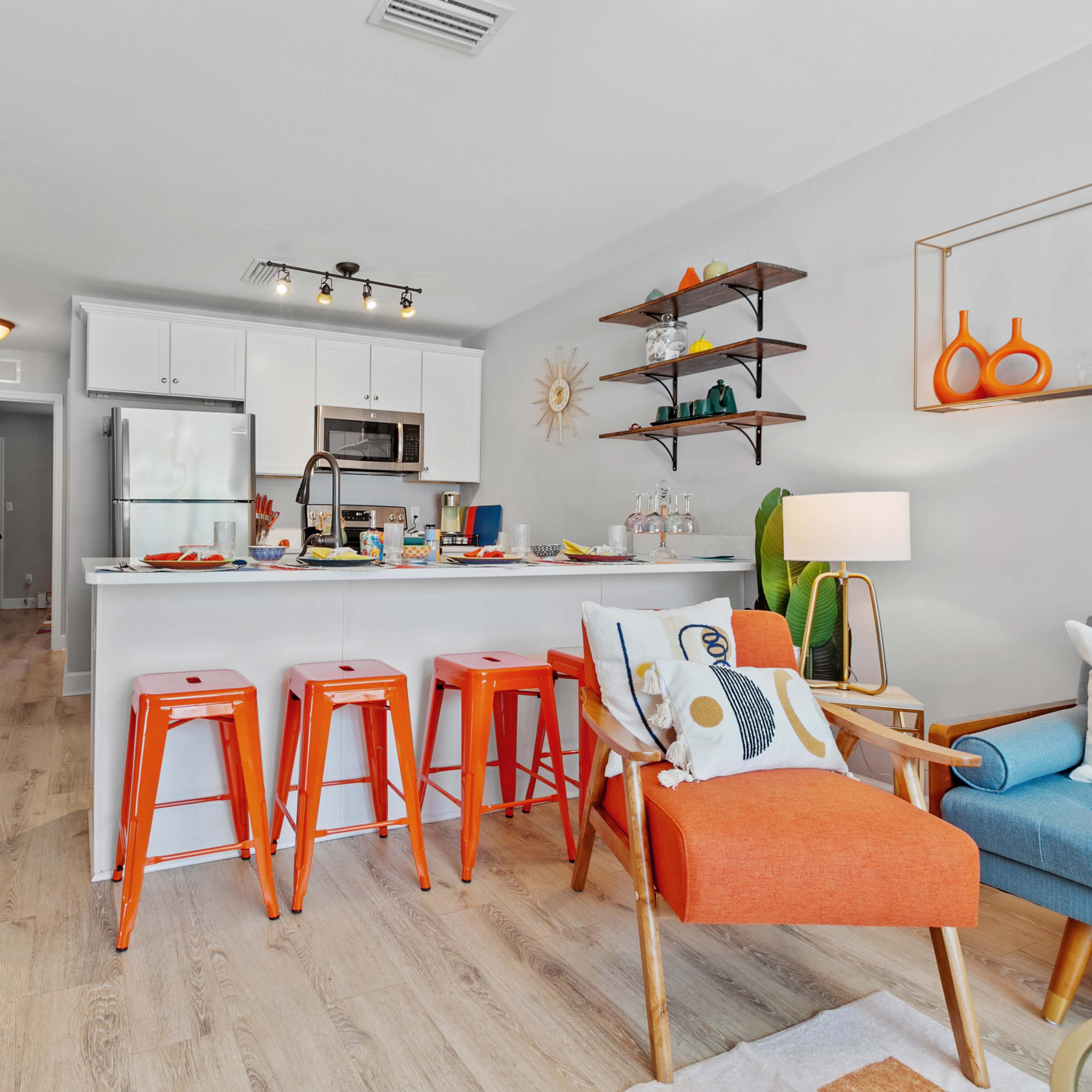 A modern kitchen and living area featuring a bar with orange stools, a well-lit space, and decorative elements on shelves.