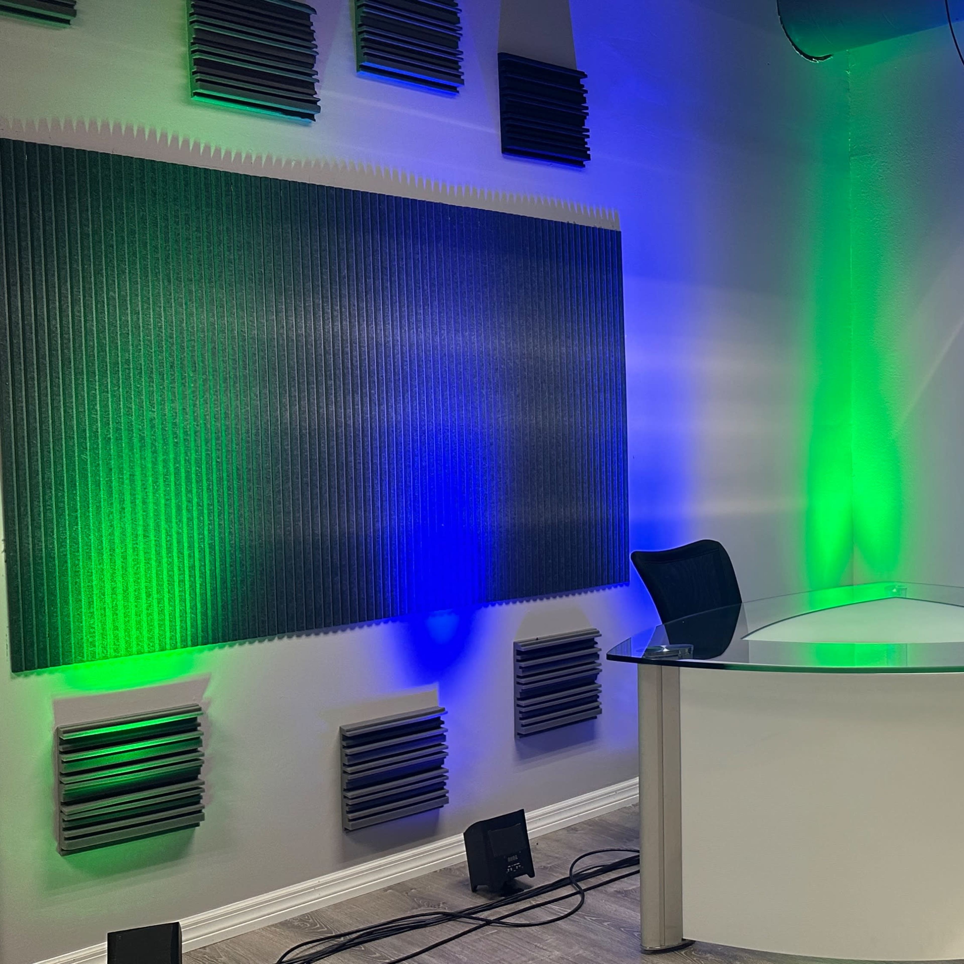 A modern, minimalistic studio with a glass desk and two black chairs, surrounded by wall-mounted acoustic panels and illuminated by green and blue lighting.