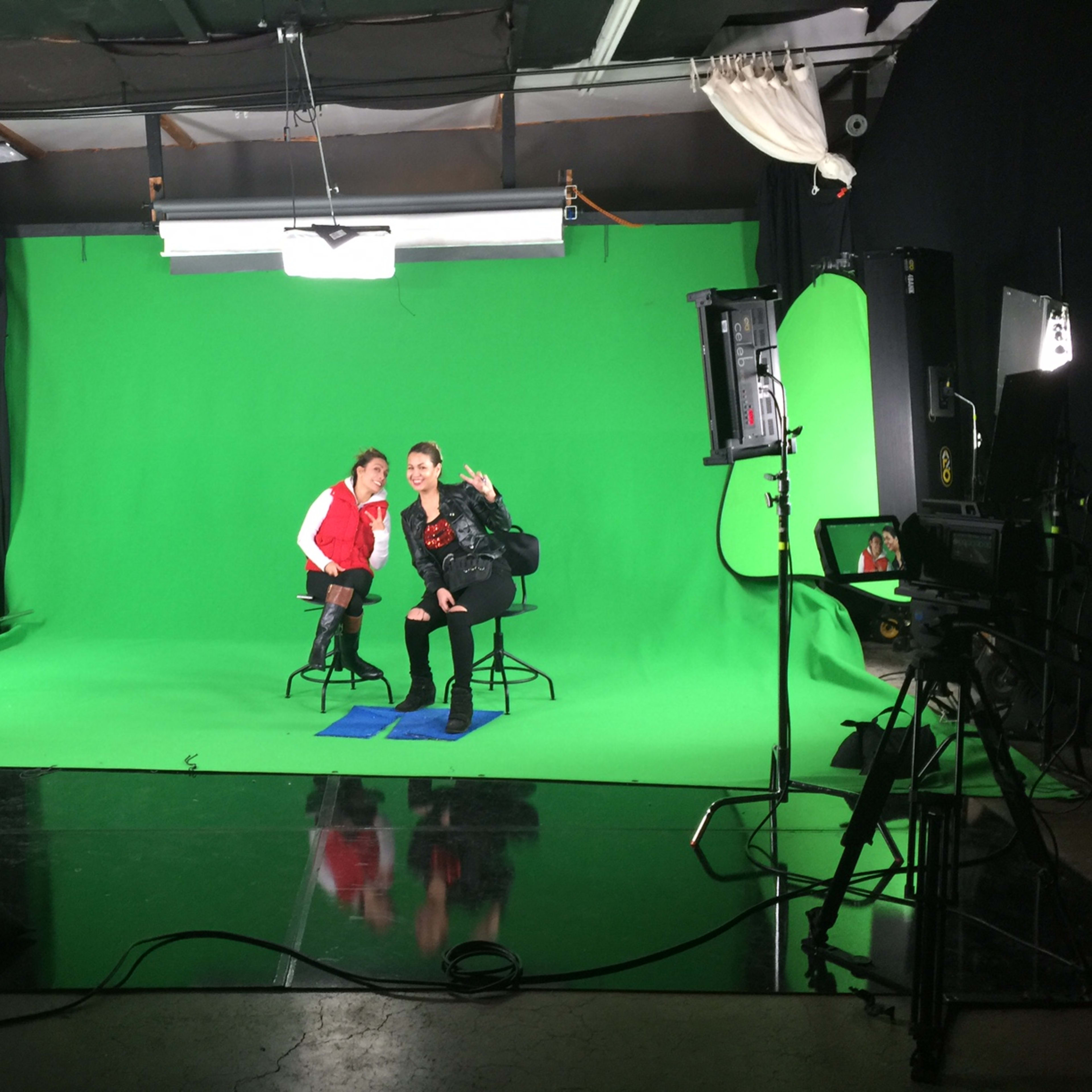 Two individuals are seated on high stools in a studio with a green screen backdrop, surrounded by lighting equipment.
