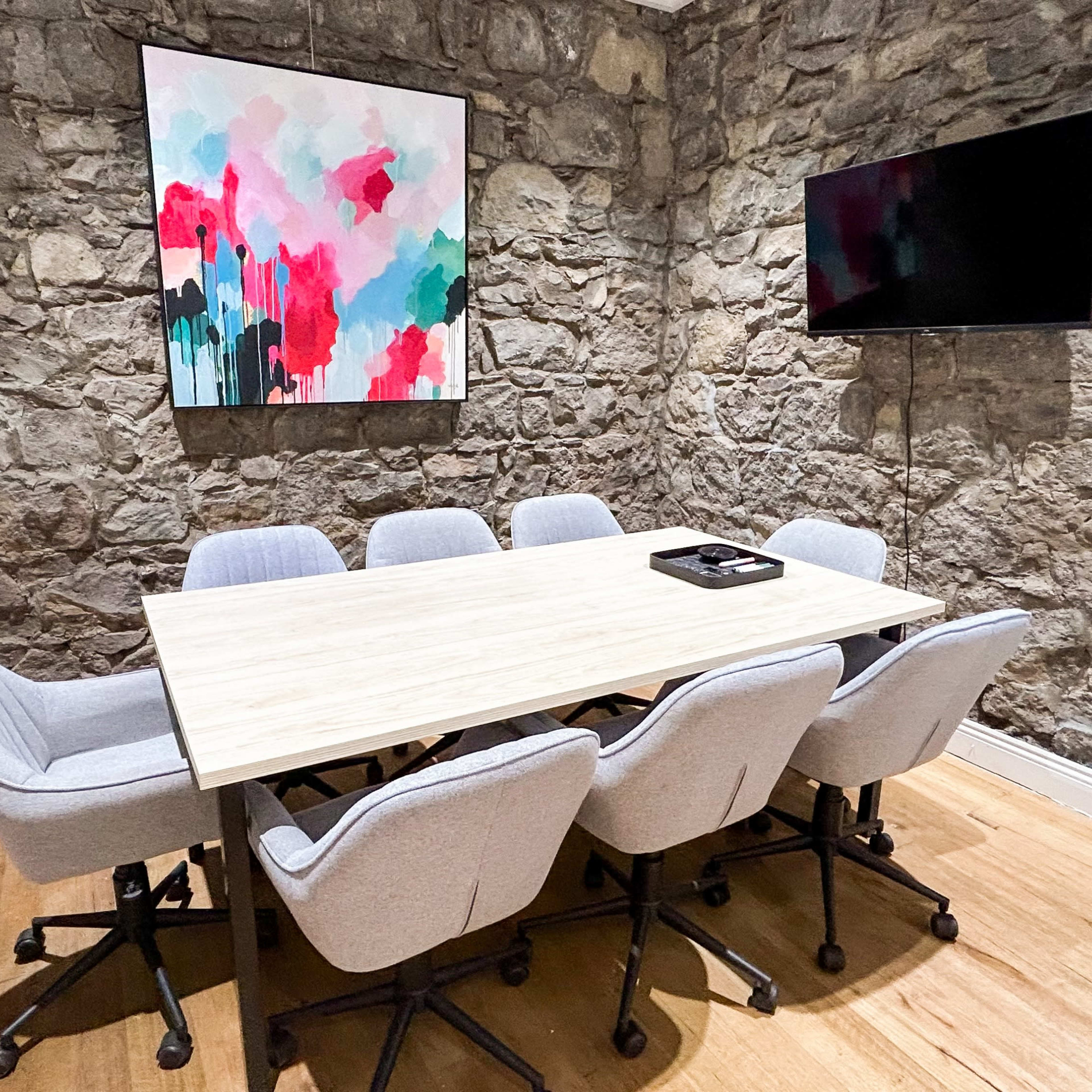 A conference room features a wooden table surrounded by gray chairs, with a colorful abstract painting on the stone wall and a wall-mounted TV.