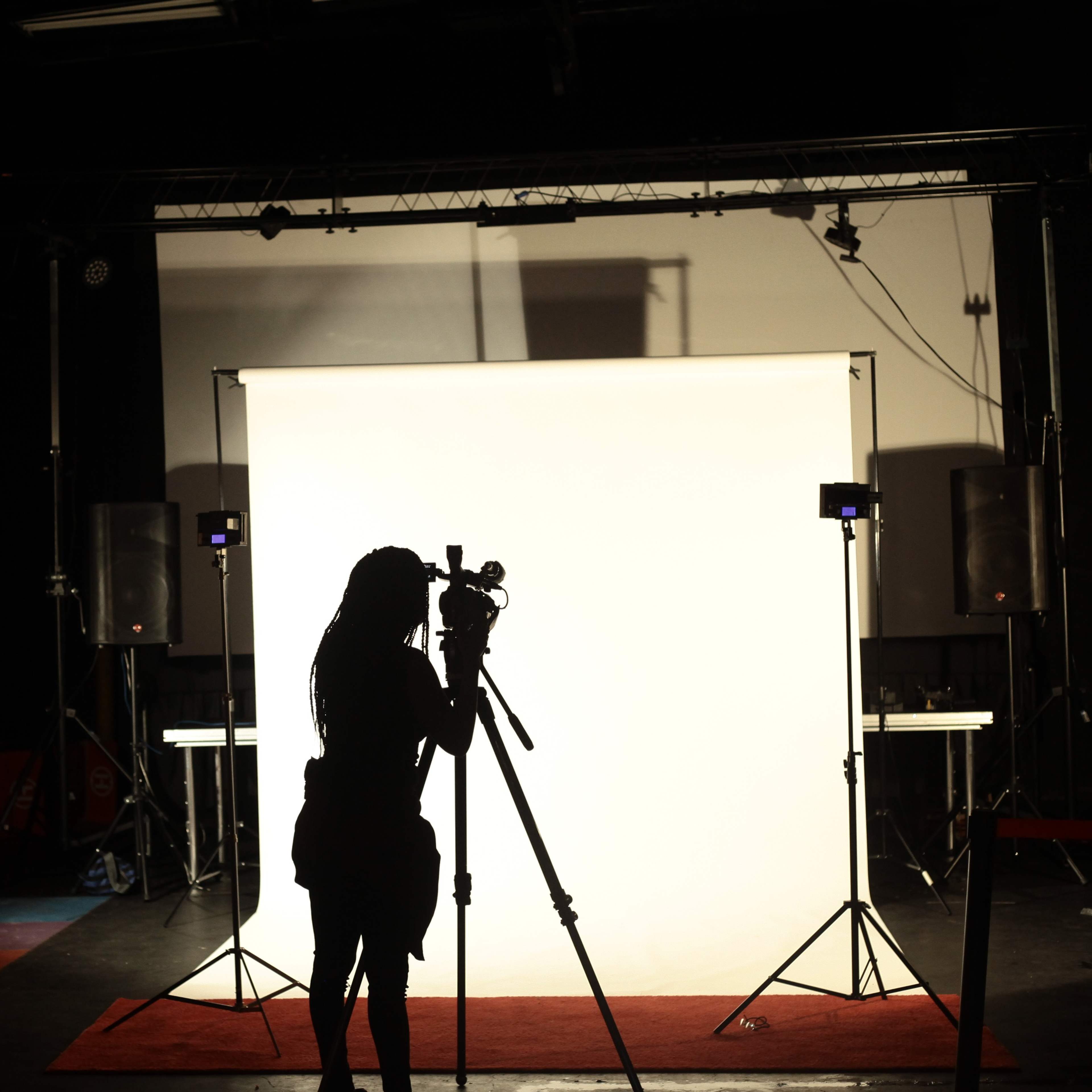 A silhouette of a person stands with a camera in front of a lit white backdrop in a photography studio.