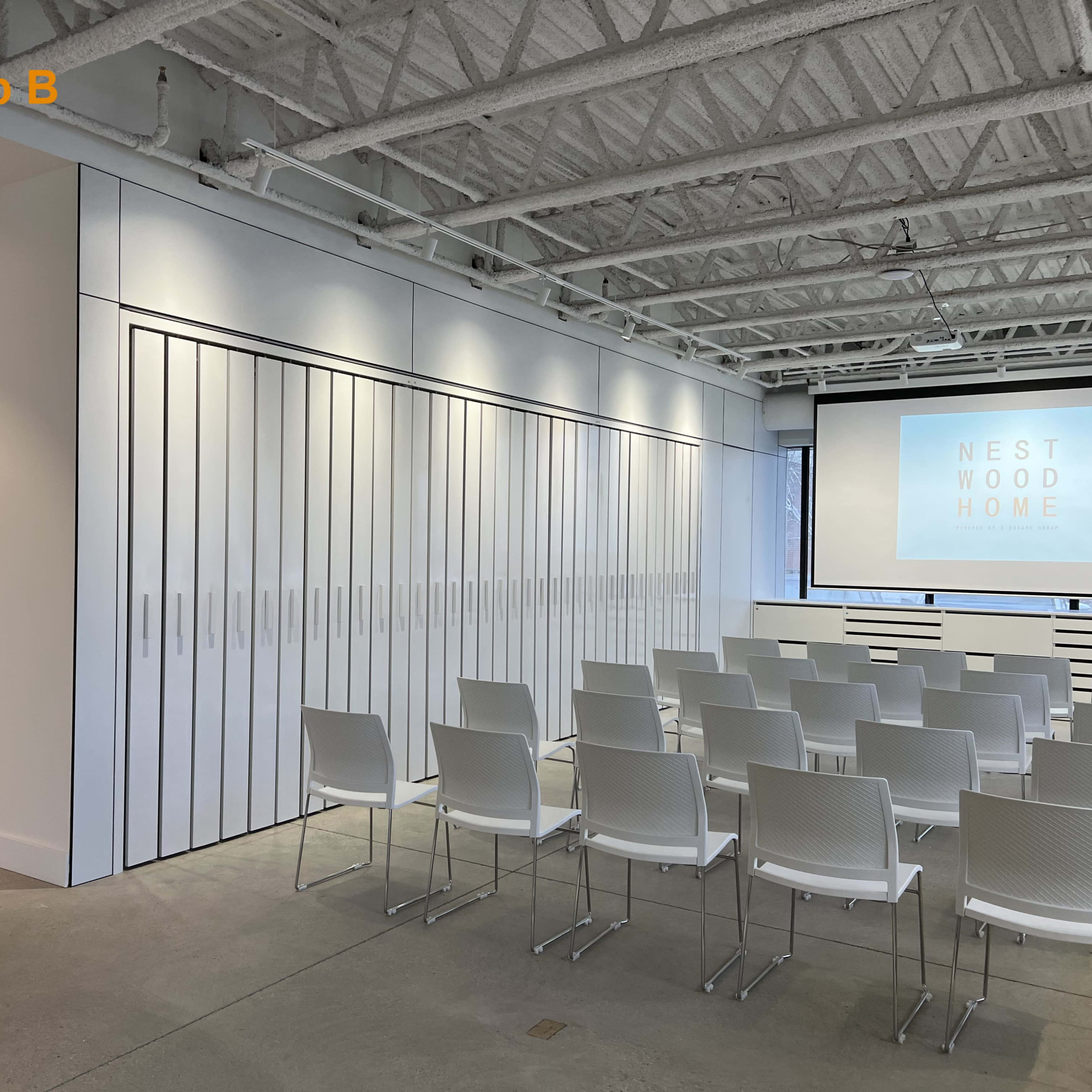 A modern workshop space features rows of white chairs facing a large screen, with white cabinetry lining one wall.