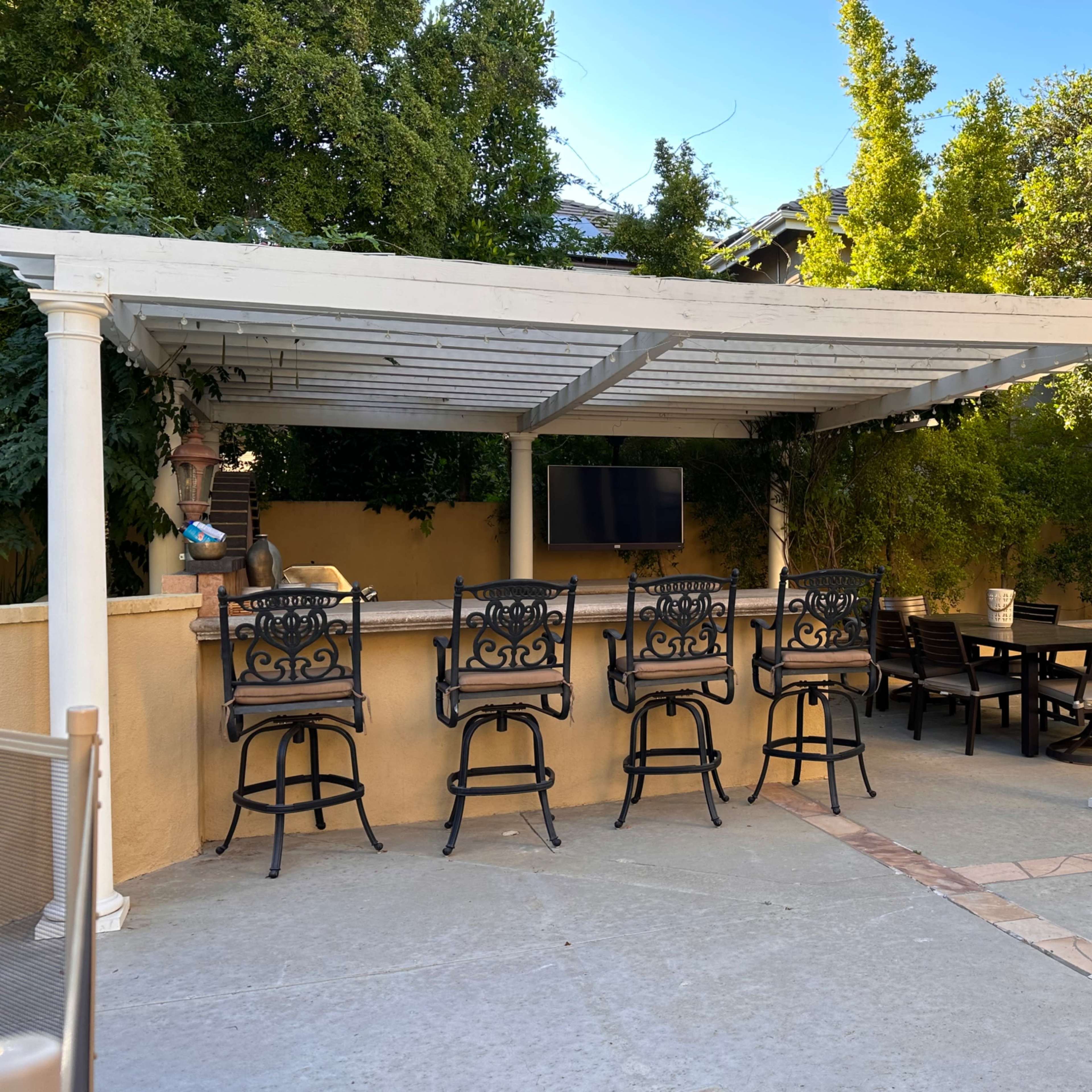 A covered outdoor seating area features several metal bar chairs and a dining table beside a wall-mounted television.