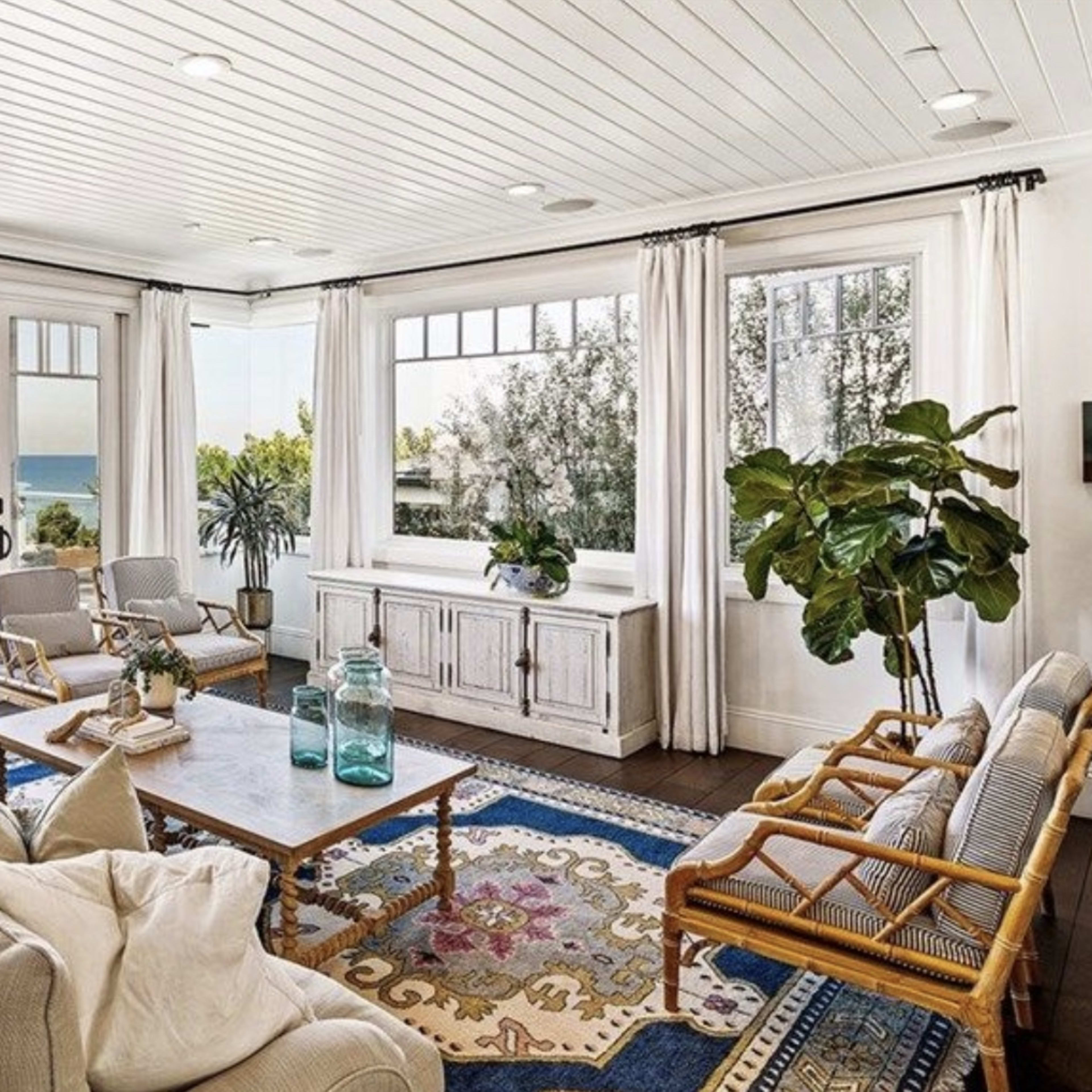 A modern living room with large windows overlooking the ocean, furnished with a light sofa, wooden chairs, and a large area rug.