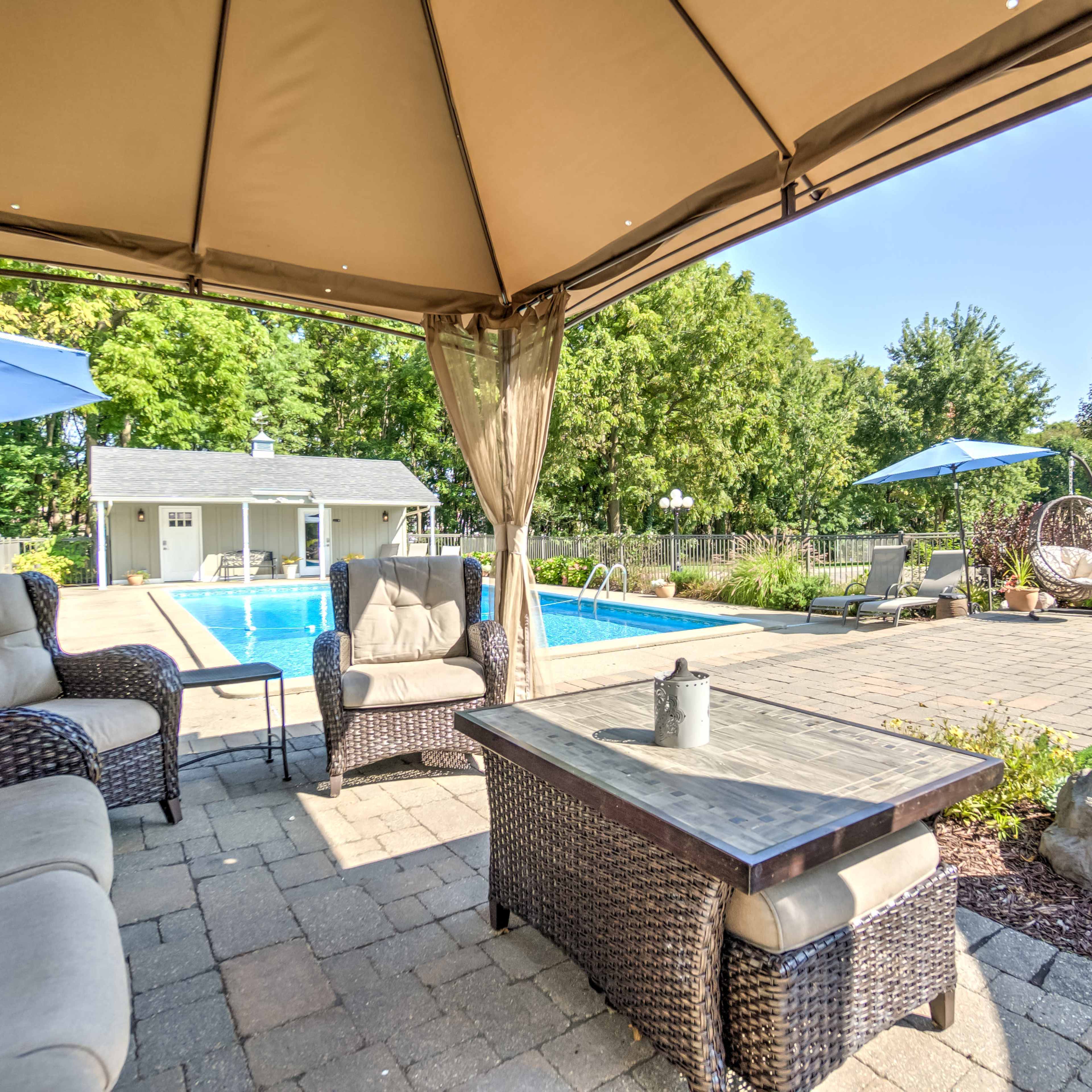 A shaded seating area with wicker furniture overlooks a swimming pool surrounded by greenery and lounge chairs.