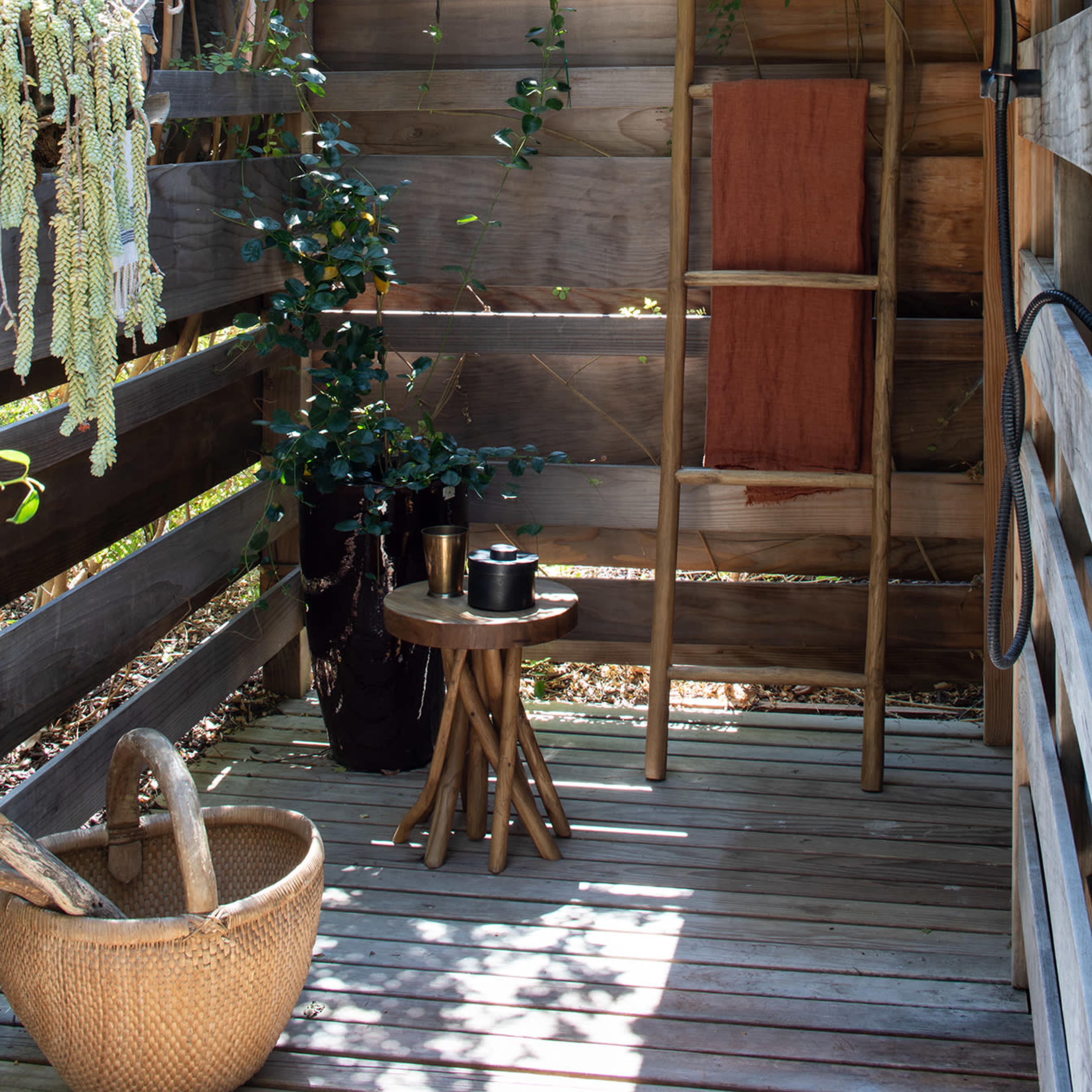 A wooden outdoor shower area features a bamboo ladder with a towel, a small wooden table, and plants along the walls.