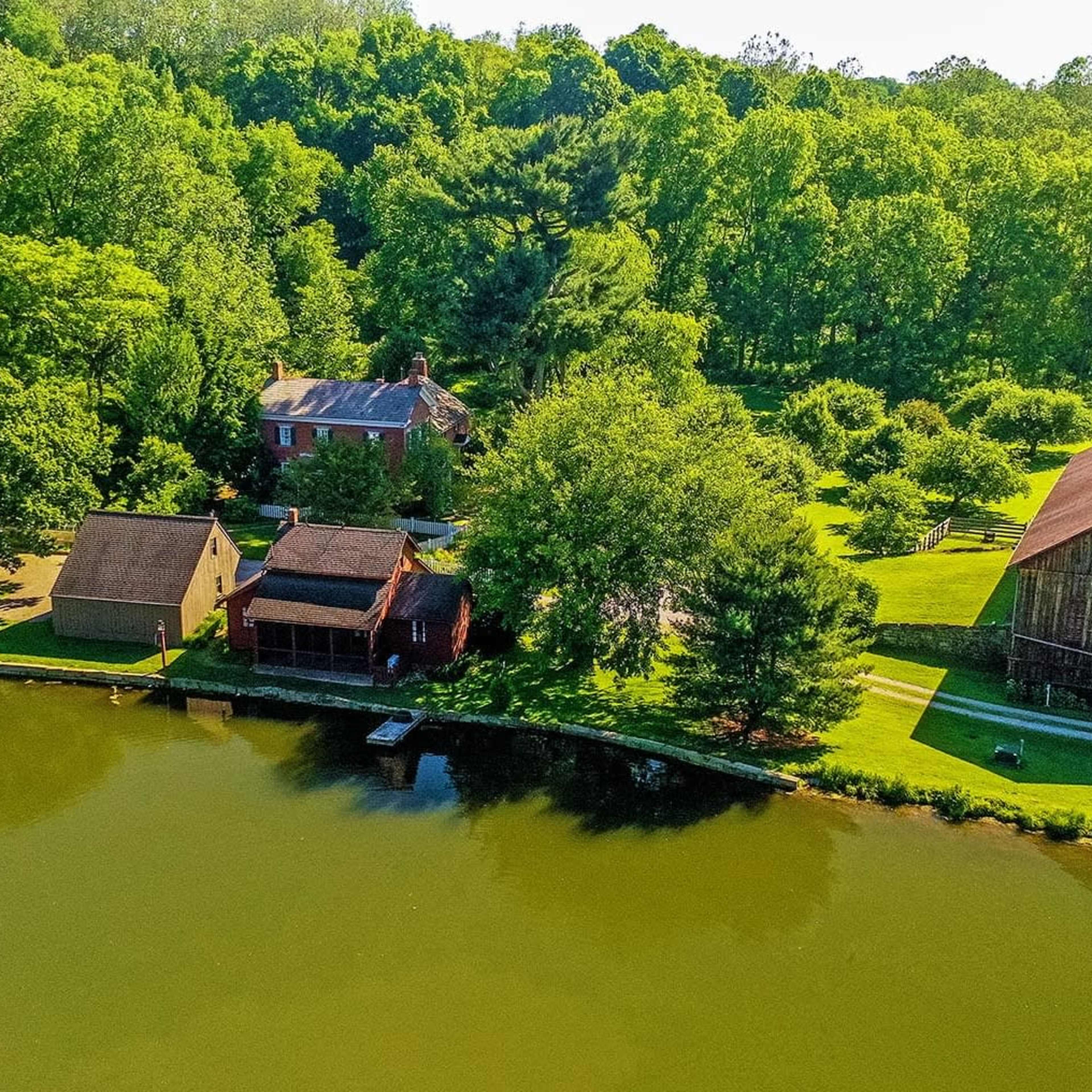 A rural landscape featuring several buildings, including a house and a barn, along the edge of a calm pond surrounded by green trees.