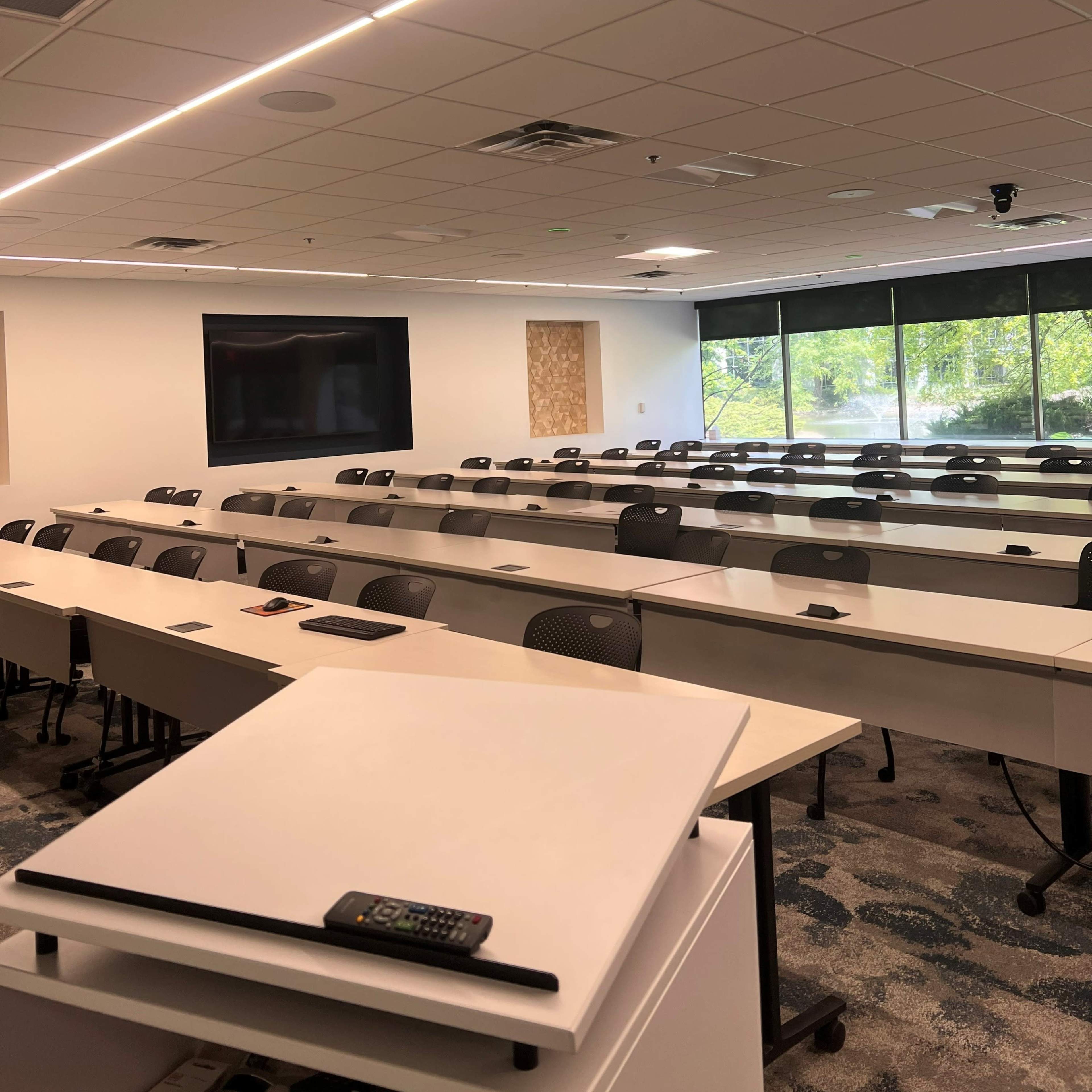A modern classroom with rows of white tables and chairs, large windows letting in natural light, and a screen mounted on the wall.