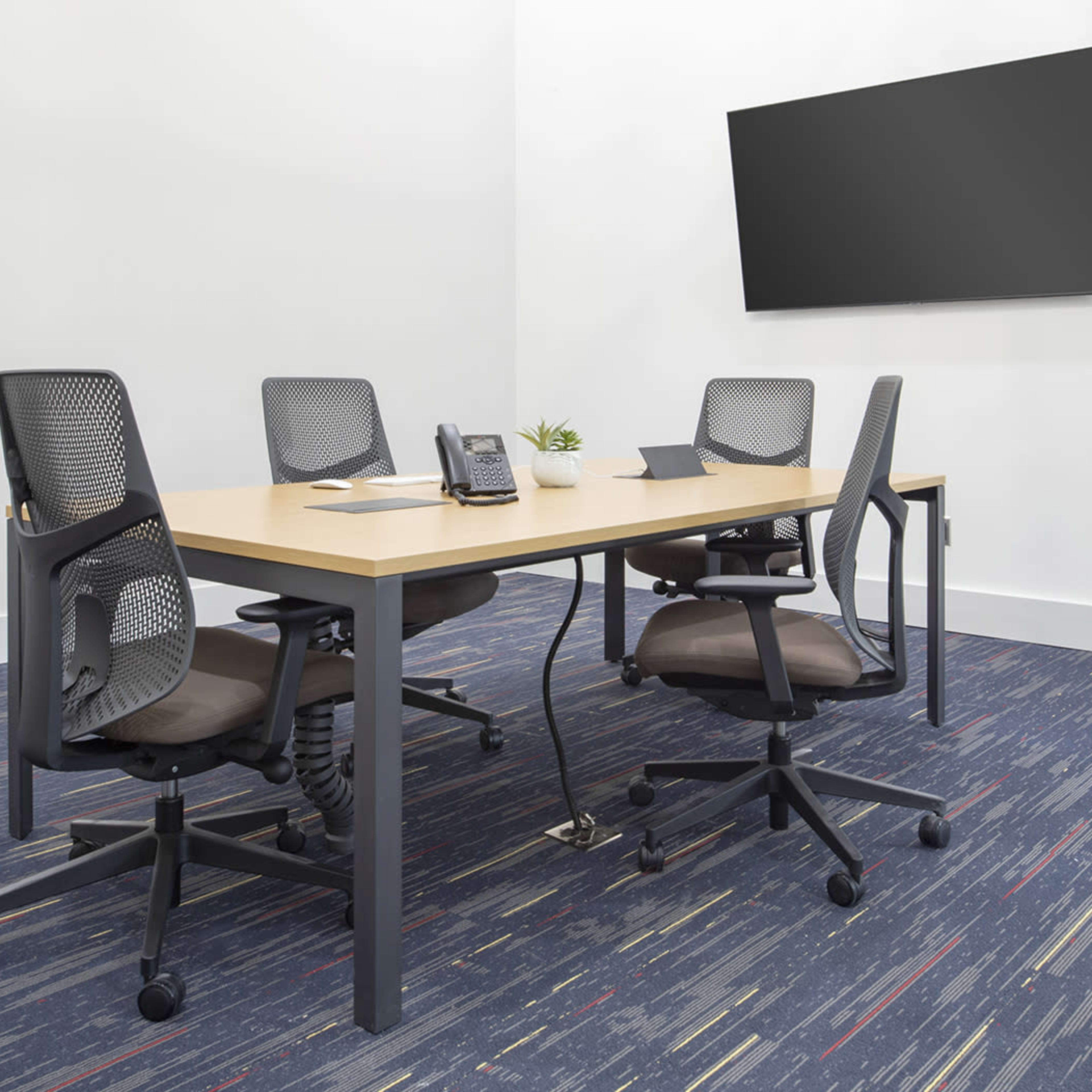 A conference room features a wooden table with four adjustable chairs, a telephone, a laptop, and a blank wall-mounted screen.