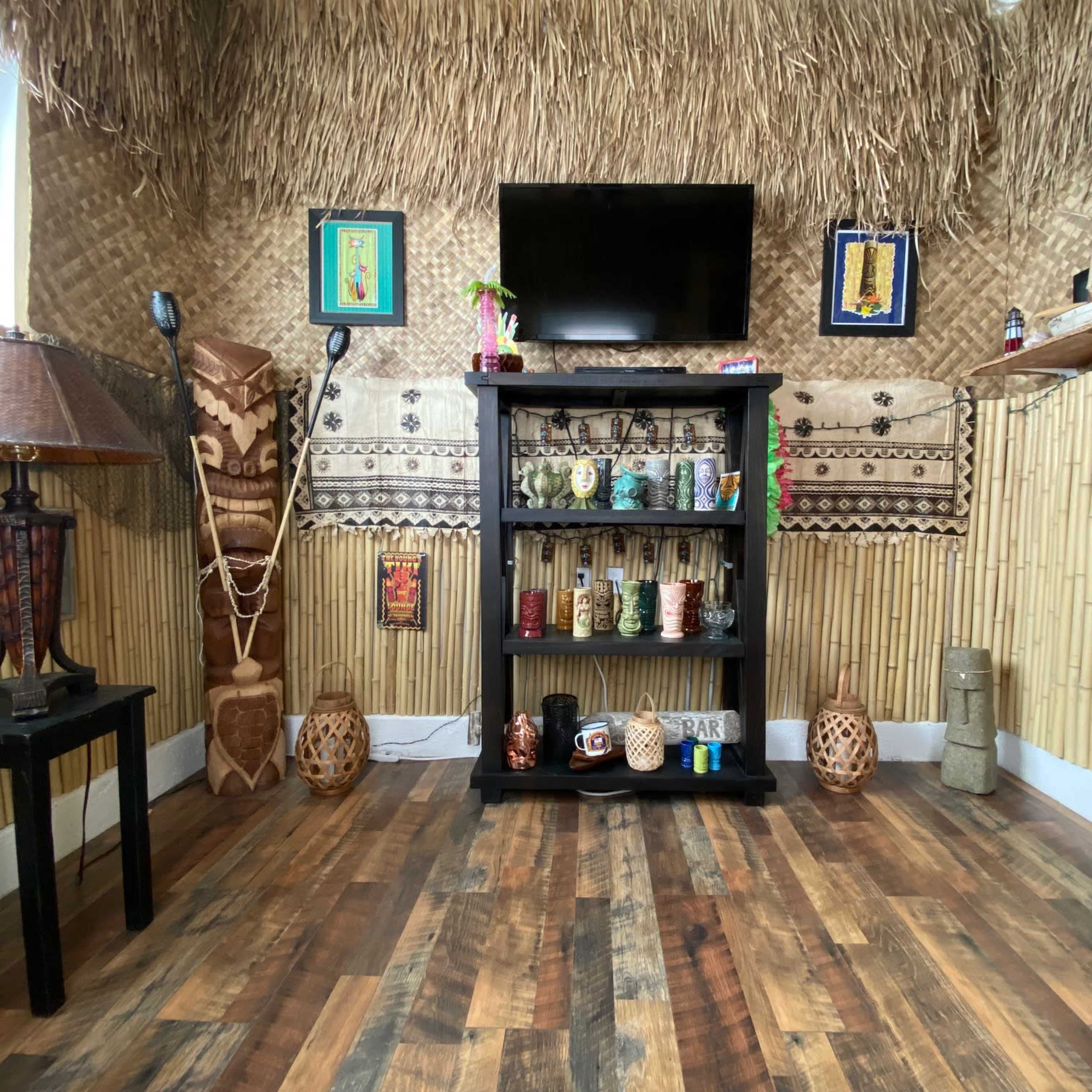 The room features a thatched wall design, a black shelf filled with colorful cups, a television mounted above it, and various decorative items scattered throughout.
